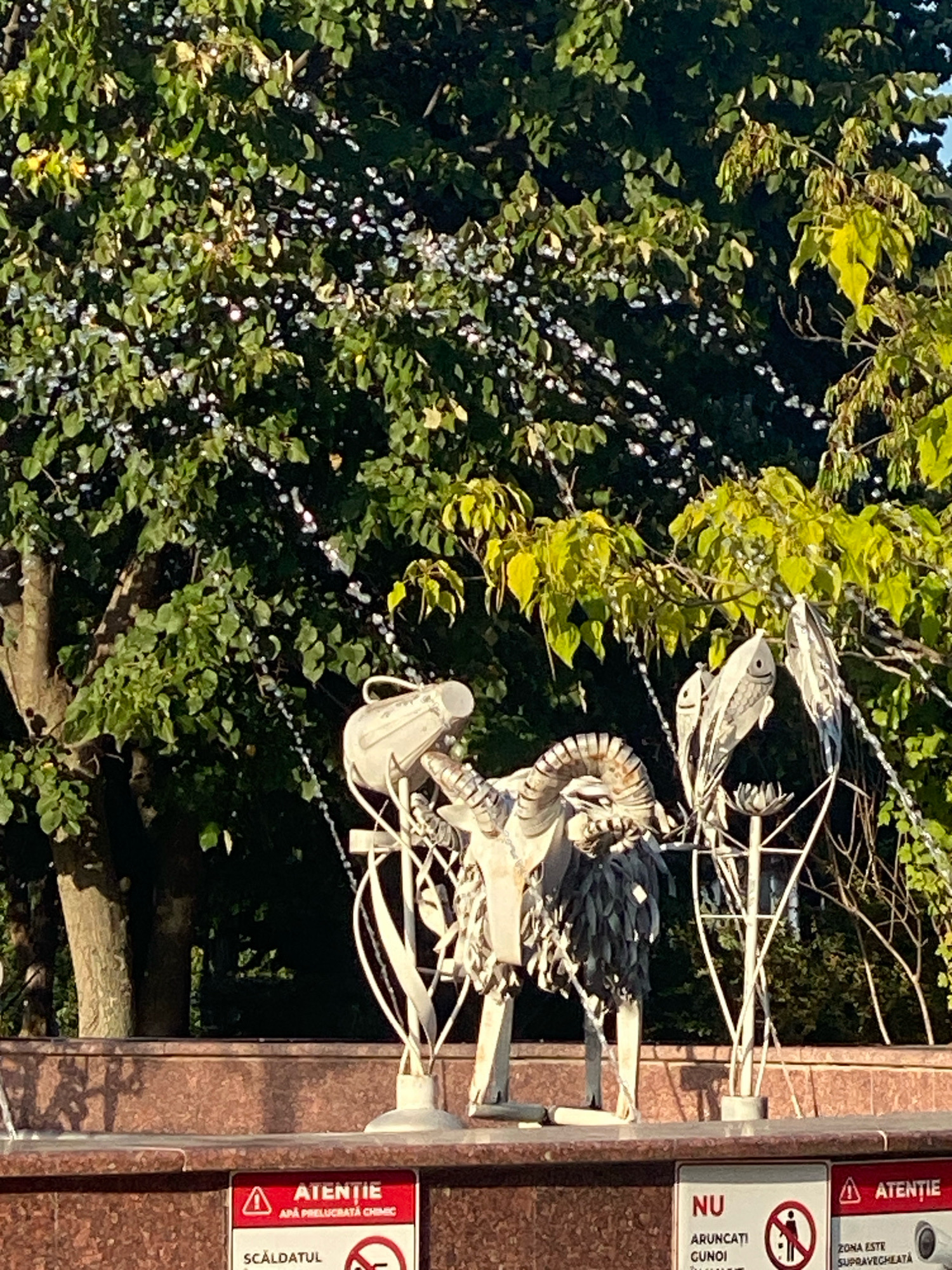 This image depicts a metal sculpture of a goat in a fountain setting, with trees and foliage in the background. The sculpture is made of various metallic elements, including what appears to be a pitcher-like object and decorative fish. Water jets are visible, indicating the fountain is operational. In the foreground, there are two signs. One sign, partially visible, is red and white and reads "ATENȚIE" and "APA PRELUCRATĂ CHIMIC" (Warning: Chemically treated water) and "SCALDATTUL" (Swimming). Another sign to the right also reads "ATENȚIE" and indicates "ZONA ESTE SUPRAVEGHEATĂ" (The area is monitored), with a security camera icon. The overall scene suggests an outdoor public space, possibly a park or square. The presence of Romanian text on the signs places the location in either Romania or the Republic of Moldova. Given the context of public fountains with specific signage, this is likely a public space in Chișinău, Moldova, a city known for its parks and fountains.