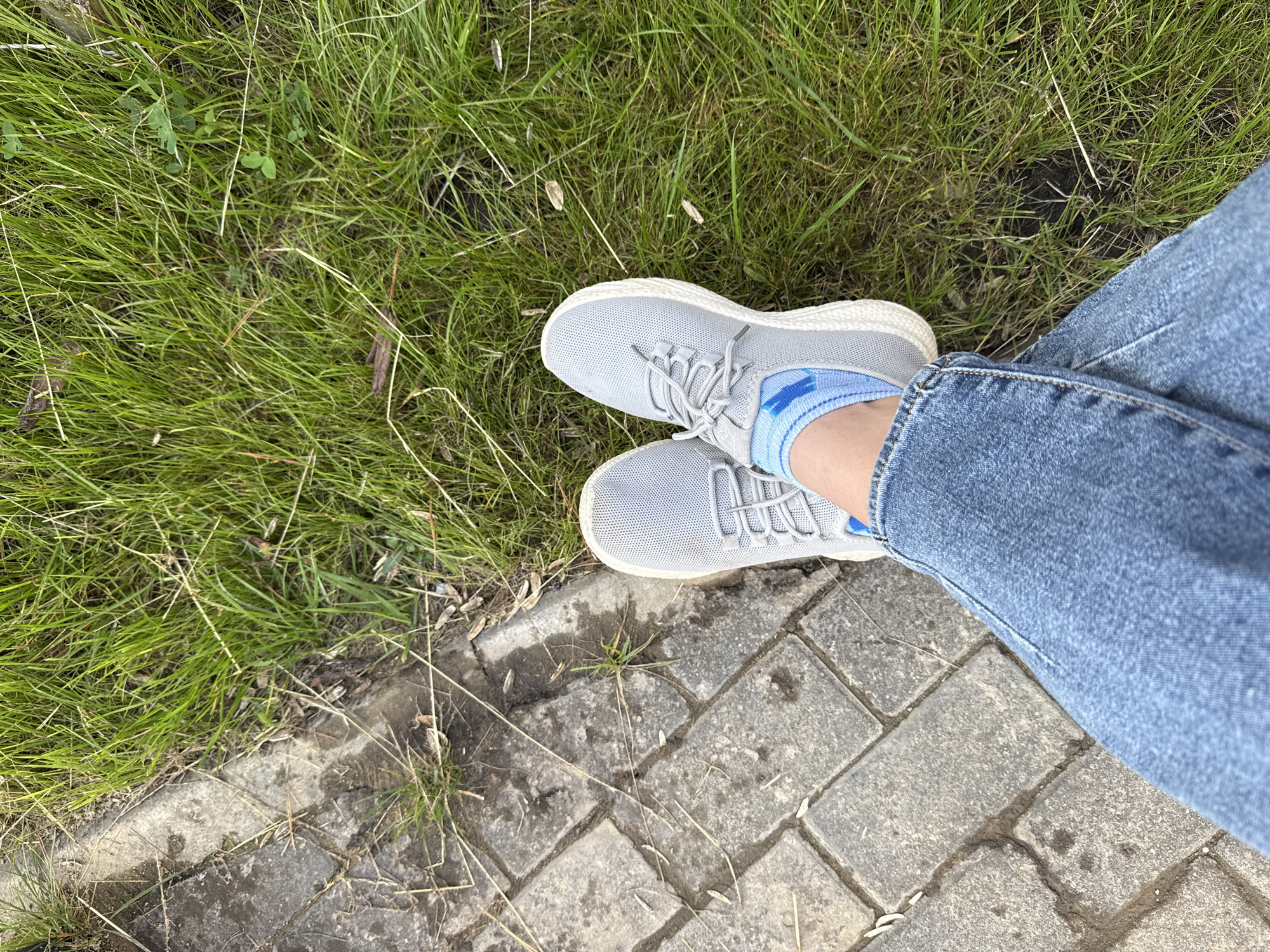 The image shows a pair of feet wearing gray sneakers and blue socks with white and blue stripes, standing on a paved walkway next to a grassy area. The person is wearing blue jeans. The scene appears to be outdoors, possibly in a park or along a street. There are no visible landmarks or identifying features that would suggest a specific location in Kazan, Russia. The lighting indicates it is daytime. The focus is on the lower legs and feet of the individual.