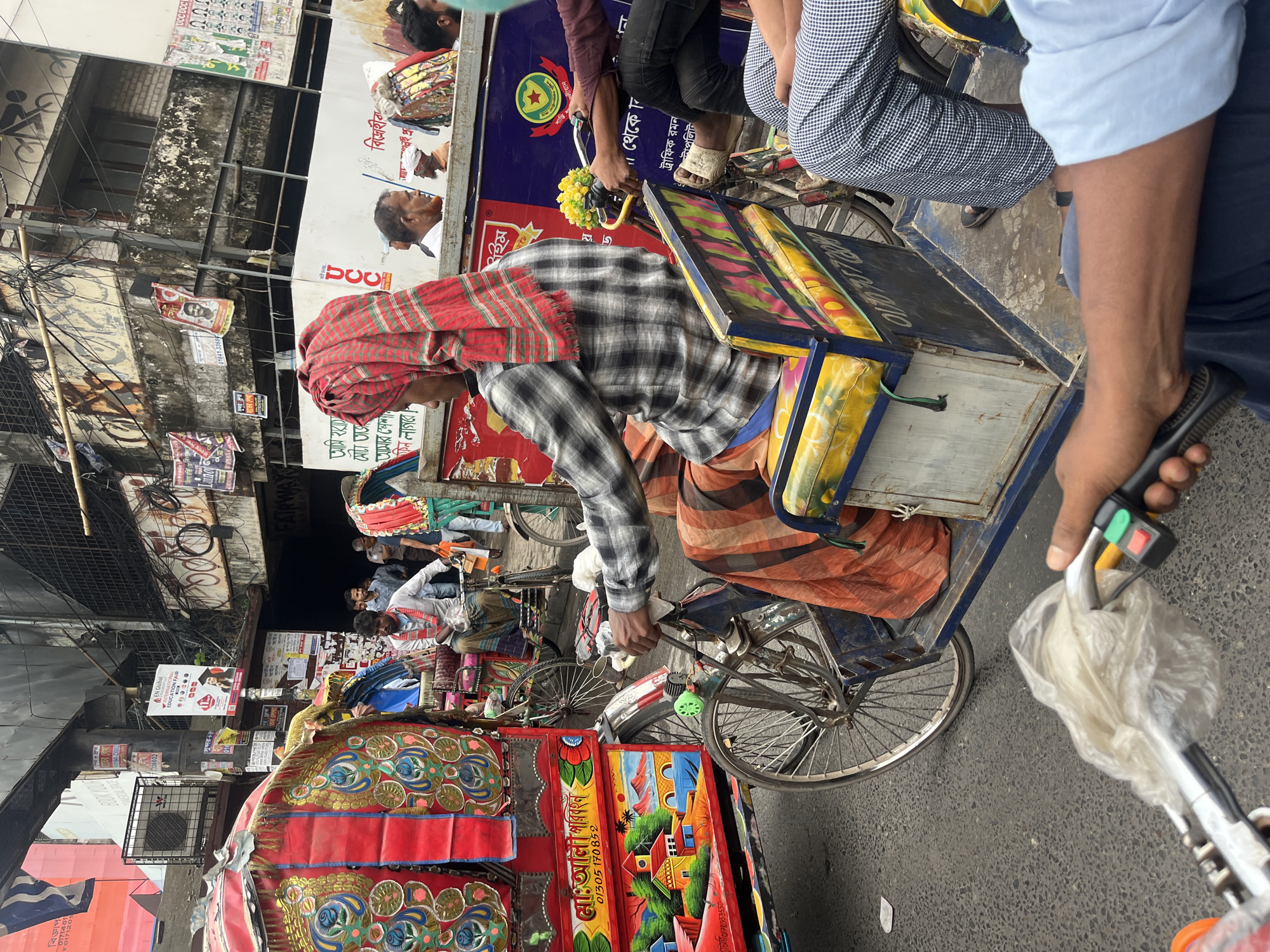 This media file captures a bustling street scene in Dhaka, Bangladesh, featuring multiple cycle rickshaws and pedestrians under daylight conditions. The image is oriented vertically and appears to be taken from a slightly elevated perspective, possibly from an adjacent vehicle or building.
In the immediate foreground, a rickshaw puller is visible, wearing a red and white checkered head wrap and a plaid shirt, actively pedaling his rickshaw. The rickshaw's passenger seat has colorful, cushion-like coverings. A small bunch of yellow artificial flowers adorns the handlebar. To the extreme right, a hand holding a black remote or controller with green and red buttons is visible, partially obscured by a transparent plastic bag.
The street is characterized by overhead electrical wires crisscrossing in a dense network. Buildings line the street, displaying signs and posters in Bengali script. Many rickshaws, distinctively adorned with vibrant, hand-painted artwork featuring landscapes, abstract patterns, and floral designs in red, gold, blue, and green, are present in the midground and background. One prominent rickshaw design shows a village scene with houses and trees.
The scene conveys a lively, everyday urban environment. Visible text on the various signs and posters includes "EN Global INTERNATIONAL EDUCATION FAIR", "মো: আলী পরিবহন" (Md. Ali Transport), "বিজ্ঞাপ" (Advertisement), "ট্রাফিক রমন", "FAIRWAY", "DOC", along with several phone numbers and other Bengali words such as "শান্তি" (Peace) and "বিদ্রোহীর" (Rebel's). The overall impression is one of movement and typical street life in a South Asian city.