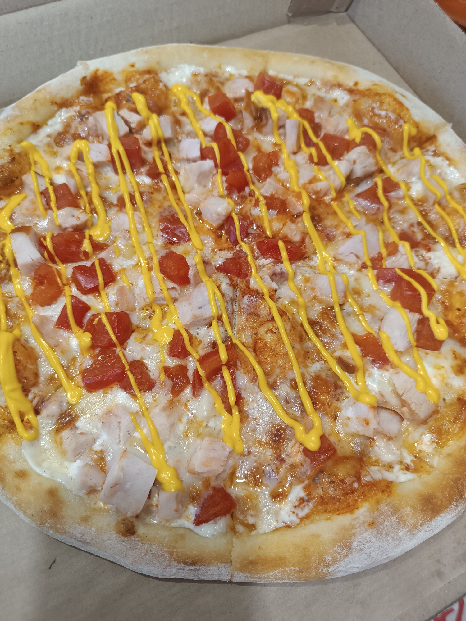 A pizza sits in a cardboard pizza box, filling the frame. The pizza has a golden-brown crust and is topped with white cheese, diced red tomatoes, pieces of cooked chicken, and a yellow sauce drizzled in a criss-cross pattern. The pizza box is open, revealing the pizza inside. The background is a brown cardboard surface. The image is taken from a top-down perspective, focusing on the pizza. The location is Chaikovsky, Russia.