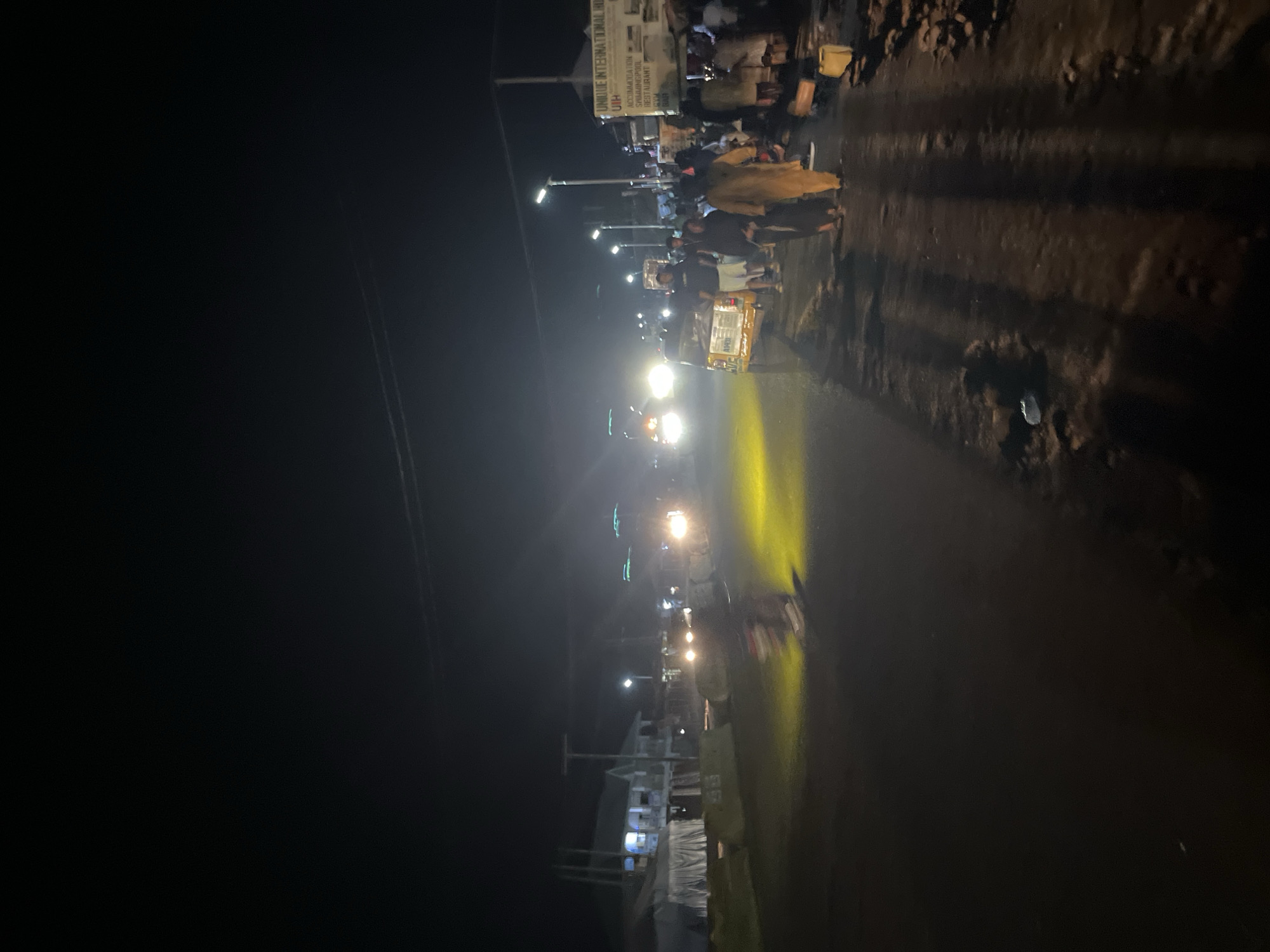 This is a vertical, low-light, night photograph of a street scene in an unidentified urban area. A yellow auto-rickshaw is parked on the right side of the road, facing away from the viewer. Several people, some blurred due to motion, are standing near the auto-rickshaw and along the right side of the road. Some are wearing traditional clothing. A sign that reads "UNIQUE INTERNATIONAL HOTEL UIH" is visible on a building to the right. Several vehicles with illuminated headlights are driving down the road, casting bright beams of light. The road is wet, reflecting the lights. In the background on the left, there are buildings with lights on, possibly shops or residences, and white tent-like structures. The overall impression is a busy street at night, with active commerce and transportation.