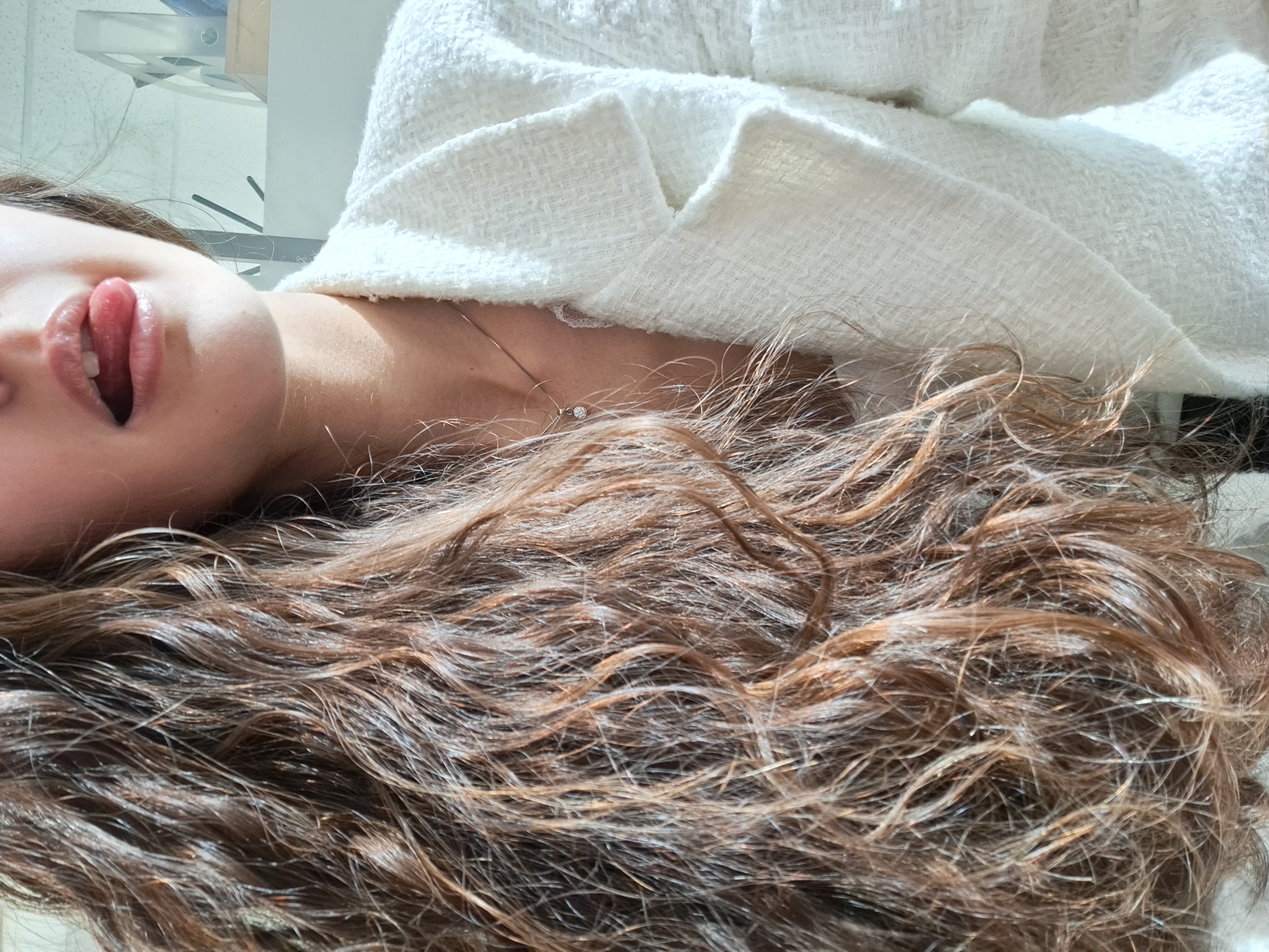 The image shows a person lying down, facing up, with their head tilted to the left. The person is sticking their tongue out. They are wearing a white textured jacket or top. A necklace with a pendant is visible on their neck. The person has long, brown curly hair that spreads out around them. The background is blurry, but appears to be a light-colored interior with some indistinct objects. The image does not provide information about Sofia, Bulgaria, or any specific location.