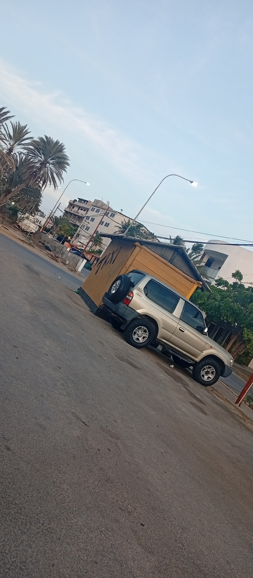 The image captures a street scene in El Yaque, Venezuela. A gold-colored Toyota Land Cruiser Prado is parked on the side of the road adjacent to a small, yellow "taxi" stand. The road surface is asphalt, showing signs of wear. In the background, there are buildings, including a multi-story hotel and various structures, indicative of a populated area. A street lamp illuminates the scene. The sky is a clear, pale blue. Palm trees are visible along the side of the road.