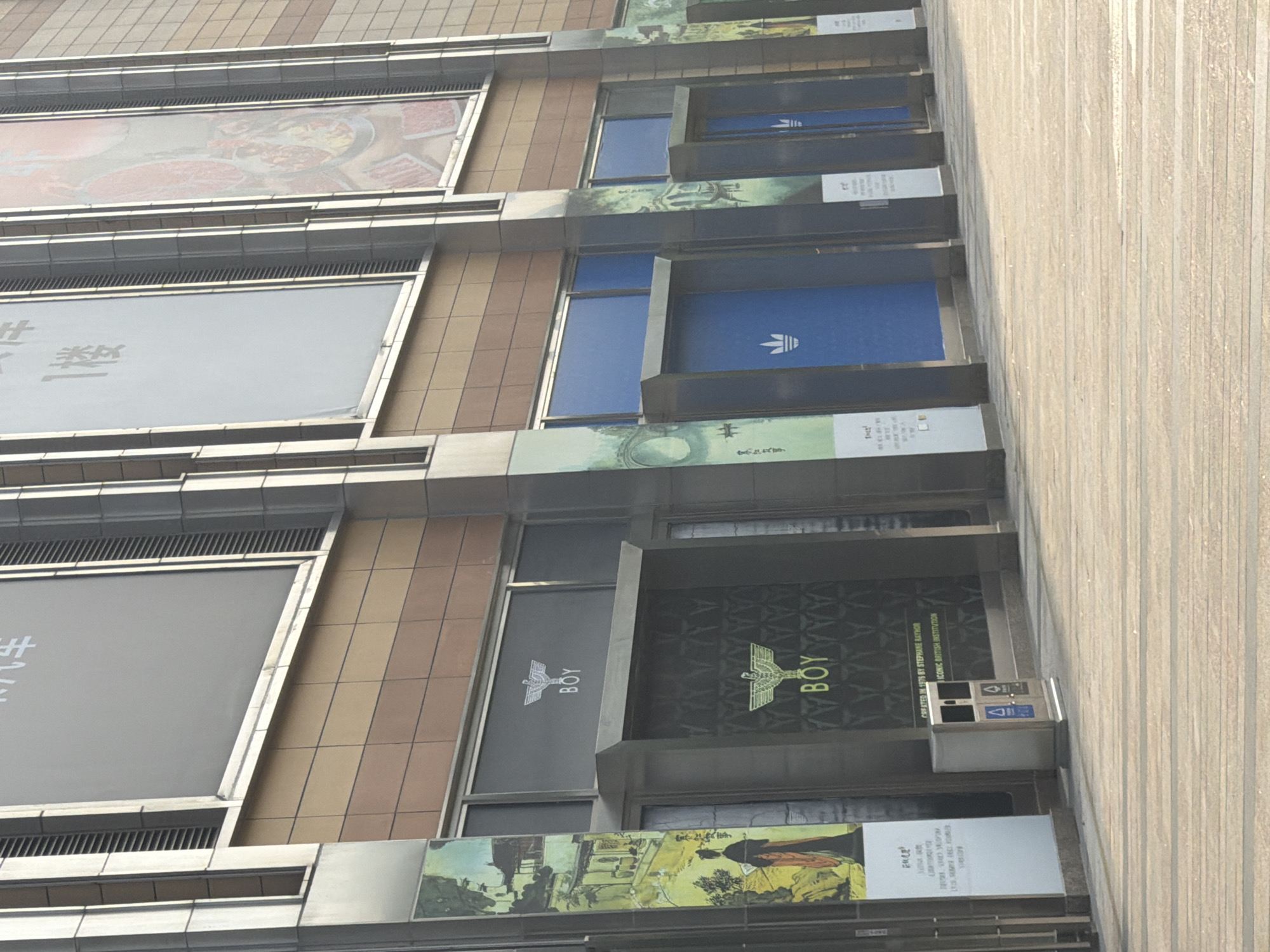 The image captures a vertical view of a building facade in Yuhuatai District, Nanjing, Jiangsu, China. The building features a series of commercial units, each identifiable by large signs. The top unit displays images of food, likely advertising a restaurant. The unit below contains a large blank, likely an advertisement. Below that is a unit prominently displaying the logo of Adidas, a multinational corporation that designs and manufactures athletic shoes and clothing. The next unit features the brand "Boy London," indicating a fashion retail outlet. Beneath it is another large advertisement. The architecture is characterized by a modular design, with each unit framed by metal structures, and a neutral color palette of creams, browns, and grays. The angle of the shot suggests a street-level perspective, with the building extending upwards. The sky is not visible, focusing attention on the urban environment and the building's commercial presence.