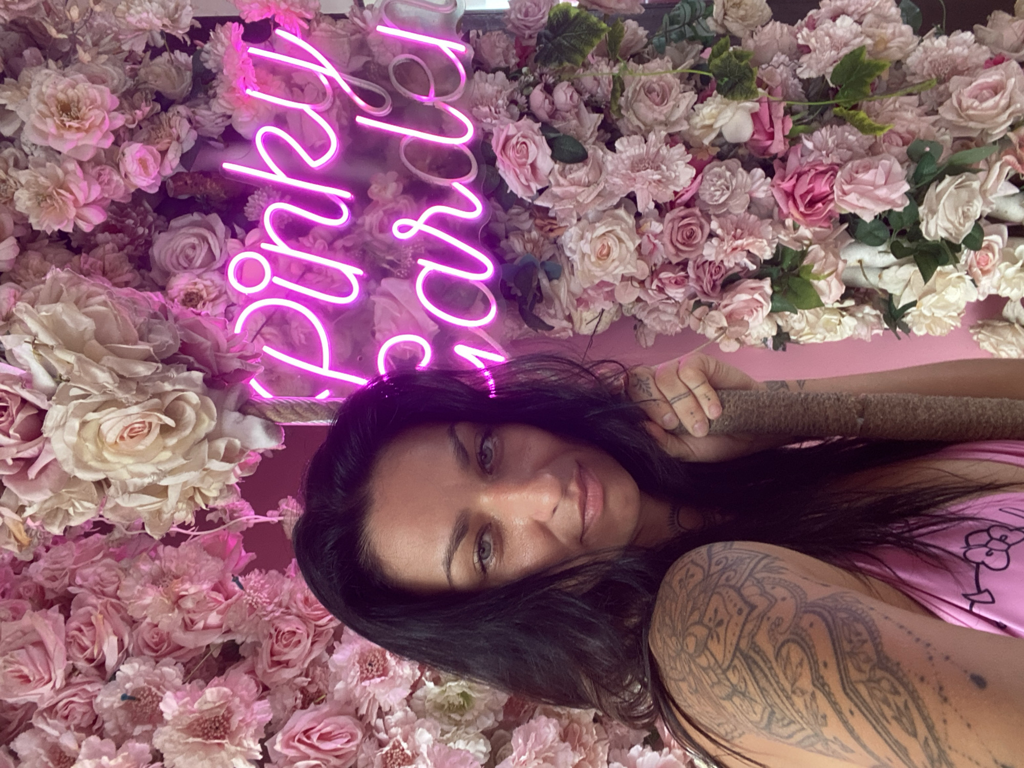 A woman poses for a photo in an indoor setting, likely a cafe or themed venue in Tien Thanh Ward, Vietnam. The image is rotated approximately 90 degrees counter-clockwise. She is positioned against a wall densely covered with artificial flowers in various shades of pink and white, including roses and carnations.
The woman has long dark hair and light-colored eyes, and she is looking directly at the camera with a soft expression. She is wearing a pink top or dress, and her left arm and shoulder are extensively covered in intricate, dark-inked tattoos featuring floral or mandala-like patterns. Her right hand is visible, holding a cylindrical, textured object, possibly a decorative element or prop.
Behind and above her, a bright pink neon sign illuminates the floral backdrop. The visible text on the neon sign clearly spells "Pinky" and partially "Garden" below it, with the full text likely being "Pinky Garden". The overall atmosphere is vibrant and decorative, characterized by the abundant flowers and glowing sign.