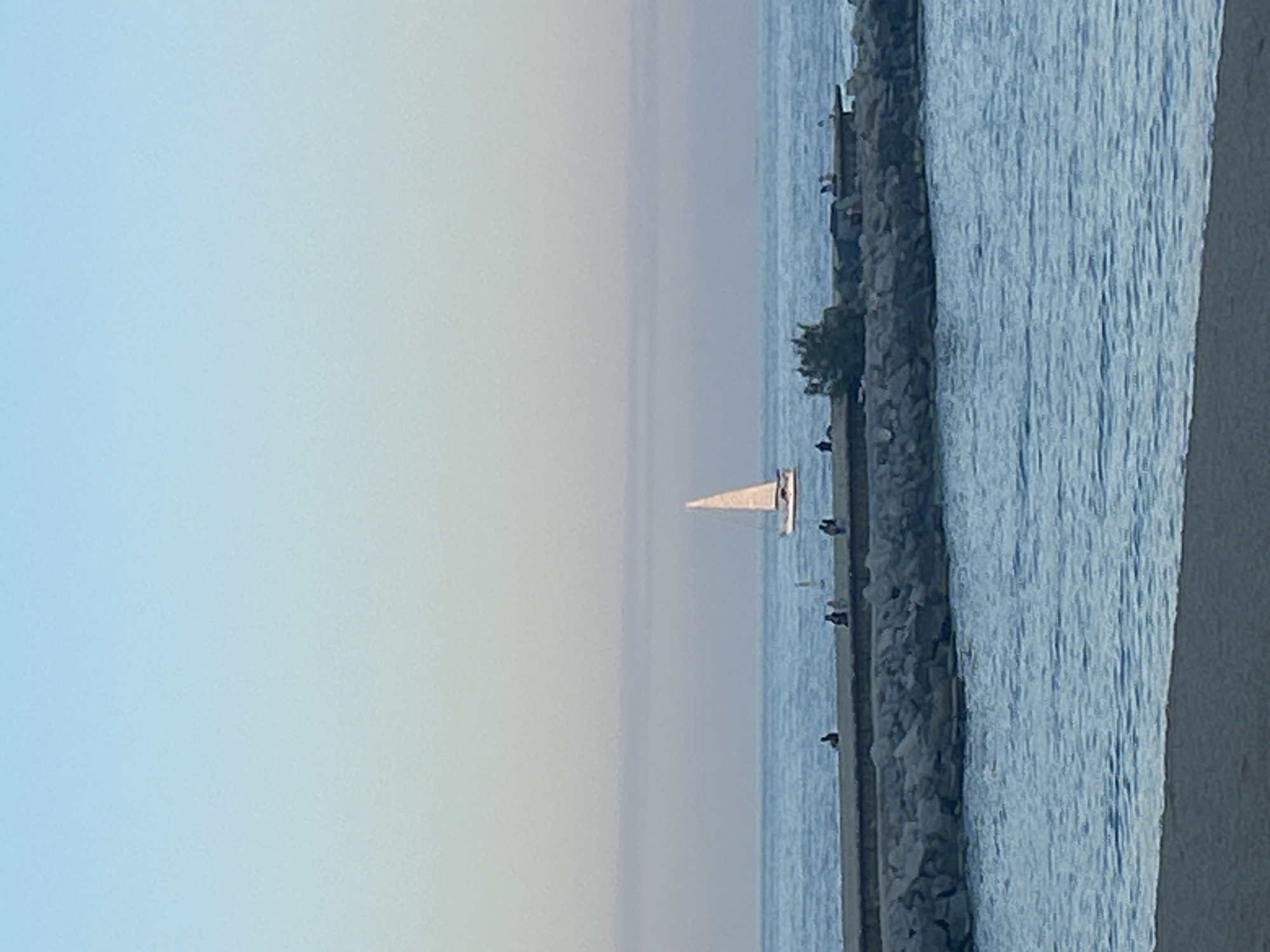 The image depicts a sailboat on the Mediterranean Sea off the coast of Barcelona, Spain. The sky is a pale blue, blending into a lighter haze over the water. The sailboat, with its white sail, is situated in the middle of the frame. A breakwater, composed of large stones, runs along the right side of the image. Small figures of people are visible along the breakwater. The water along the breakwater is a darker blue compared to the open sea. A partial view of a concrete structure is visible at the right edge.