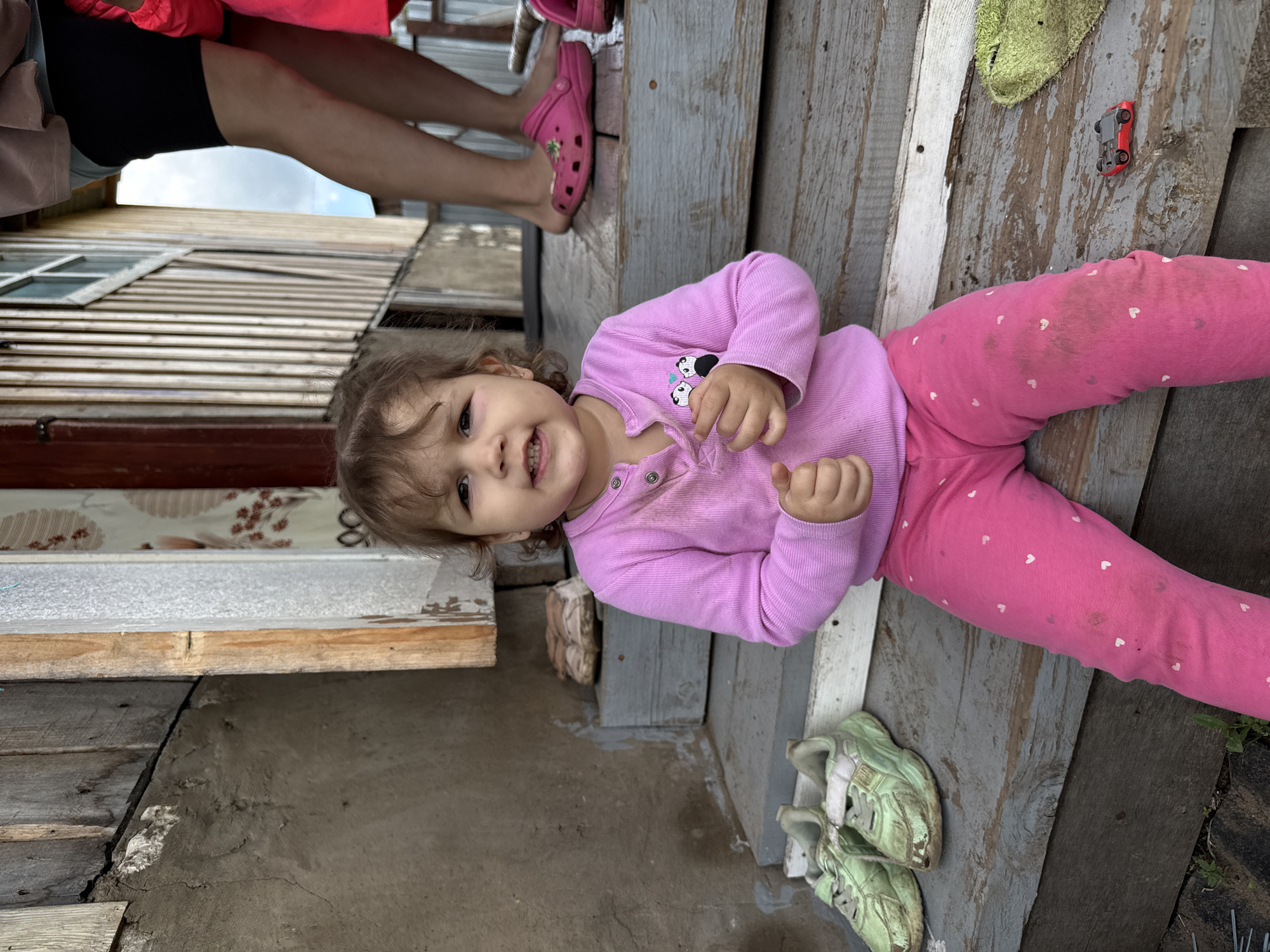 A toddler is lying on a gray wooden deck, wearing a pink long-sleeved shirt with panda bear graphics and pink pants with white hearts. There are dirt stains on her pants and shirt. The toddler is looking at the camera and smiling, with both hands raised. To the right of the toddler, on the wooden deck, are a pair of weathered green shoes, a small red toy car, and a green cloth. Above the toddler, a woman's legs and feet are visible, wearing black shorts, a pink top, and pink clogs. The edge of a building is visible to the left, with a window and wood paneling. The photo was taken outdoors in the city of Уральское, Russia.
