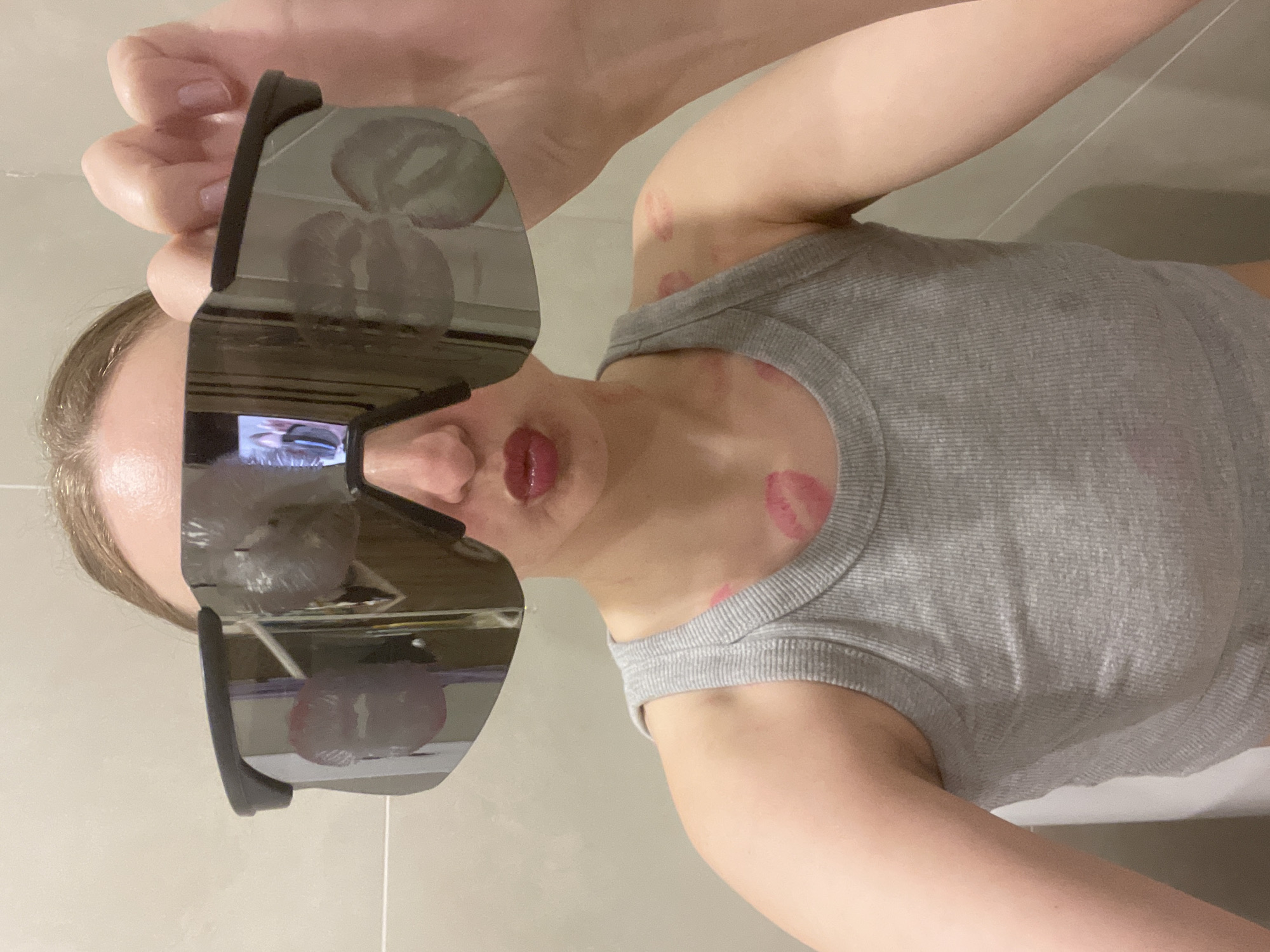 A fair-skinned woman in a grey tank top is holding large, black sunglasses up to her face. The sunglasses reflect several red lipstick marks on the lenses. Additional red lipstick marks are visible on the woman's neck and upper chest. The background appears to be a bathroom setting with light-colored tiles. No evidence suggests the image was taken in Подшивалово, Russia.