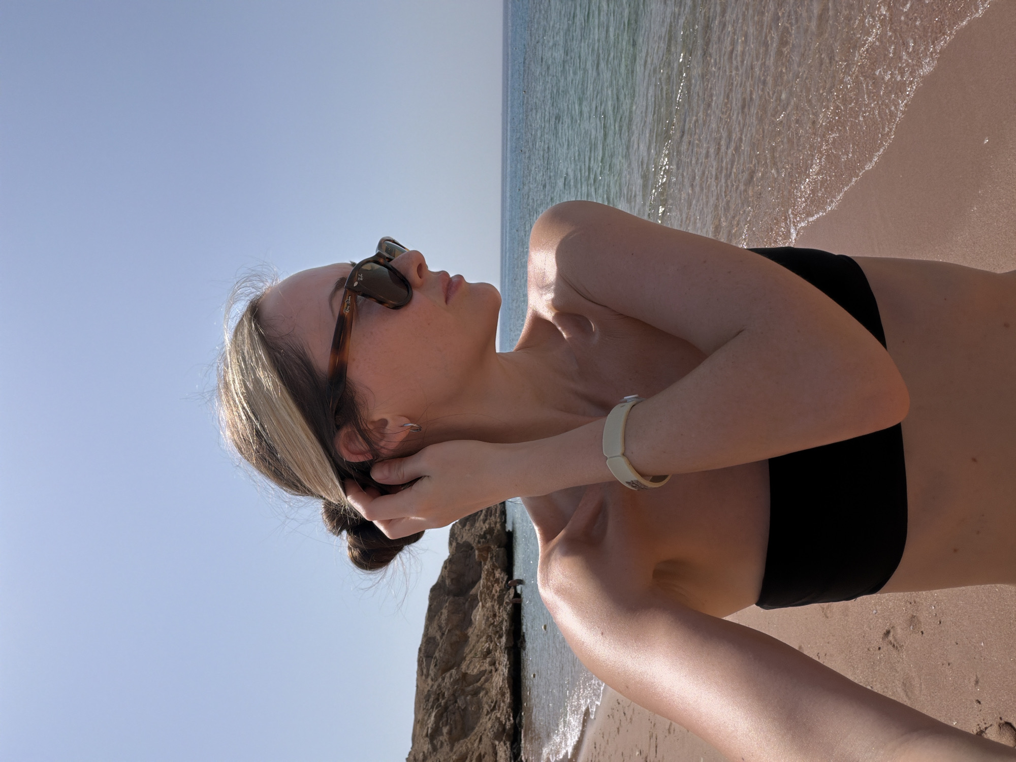This portrait-oriented image features a woman relaxing on a sunlit sandy beach under a clear blue sky. She is depicted from the chest up, reclining on her back, with her head tilted upwards, looking towards the sky. She wears dark, tortoise-shell patterned sunglasses, with the brand "Ray-Ban" visibly printed on the right temple arm. Her attire includes a black bandeau bikini top.
Her hair, which is dark with distinct blonde highlights, is pulled back into a bun. Her hands are positioned near her head, with one hand gently touching her hair while the other is near her ear. A light-colored, possibly ceramic or plastic, bracelet with a subtle pattern is worn on her left wrist.
The background shows the calm ocean to her right, with small, shimmering waves gently washing onto the light brown sand. To her left, a large, rugged rocky outcrop or cliff face rises from the beach. The sky is a cloudless, bright blue, suggesting a clear and sunny day. Strong sunlight illuminates the scene, casting highlights on her skin and hair, and creating a relaxed, summery atmosphere.