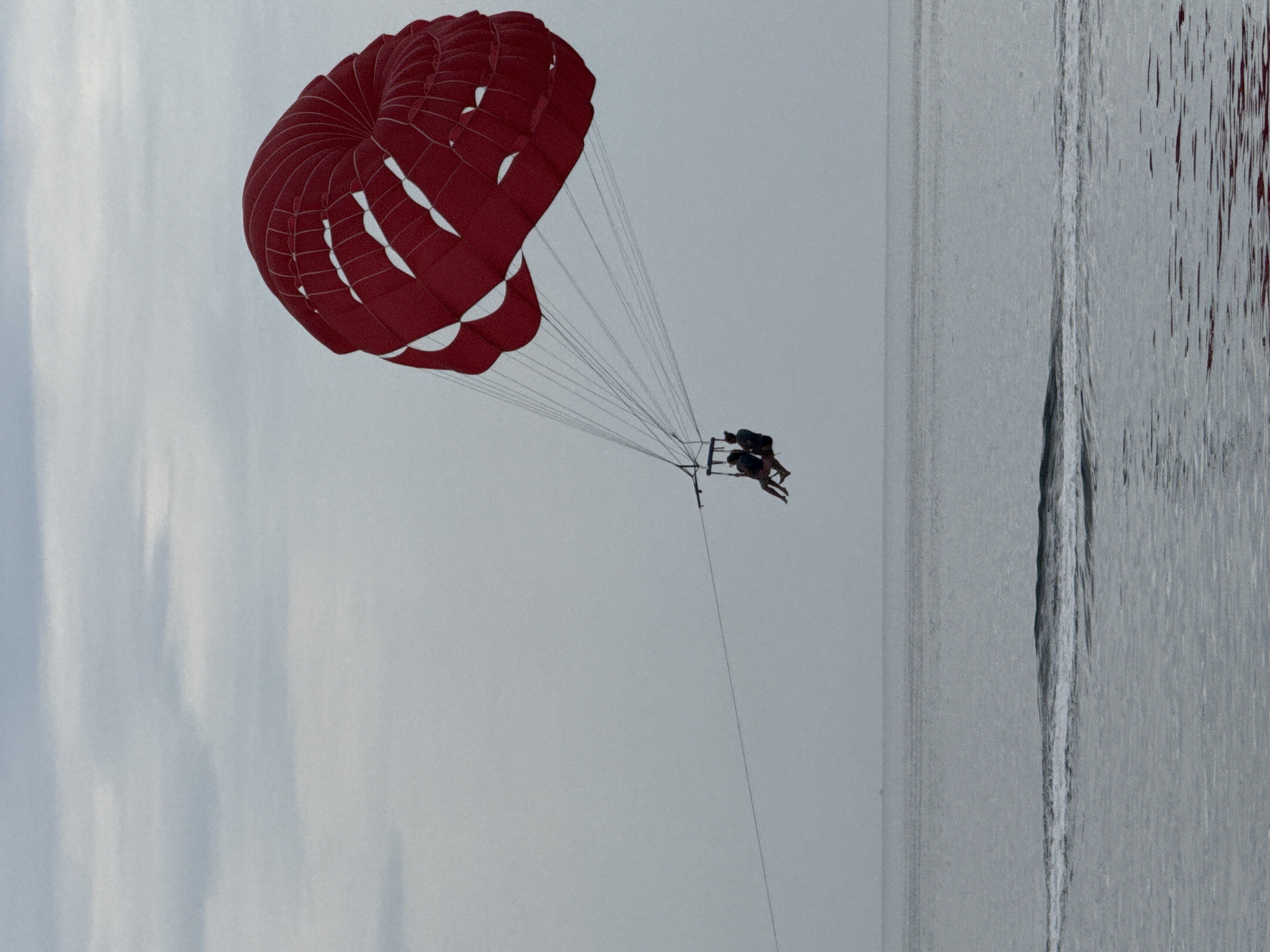A red parasail with two figures underneath hangs in the sky over the water. The location is possibly Budva, Montenegro. The water has a gray hue, indicative of possible cloud cover. The horizon is clearly visible.