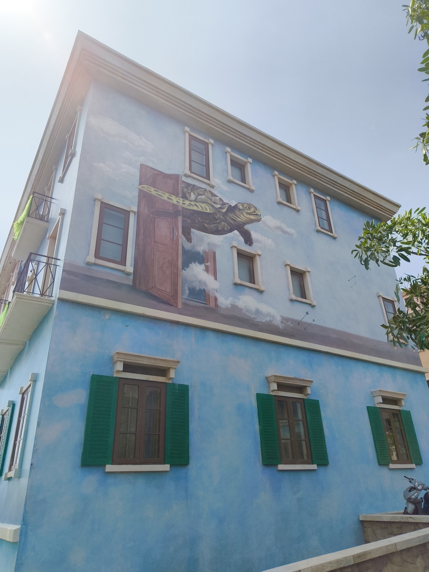 This is a medium shot, angled, exterior photograph taken during the daytime of a building in Phú Quốc, Vietnam. The building is painted a bright blue and features a large mural of a sea turtle swimming through the sky, emerging from an open doorway. The turtle mural extends across multiple stories of the building, with painted clouds and a blue sky background. The building has several windows, some with green shutters, and balconies with black railings. Greenery is visible on one of the balconies and on a tree branch extending into the frame from the right. A parked scooter is visible in the lower right corner. The sky is bright and clear with some lens flare in the upper left quadrant.
