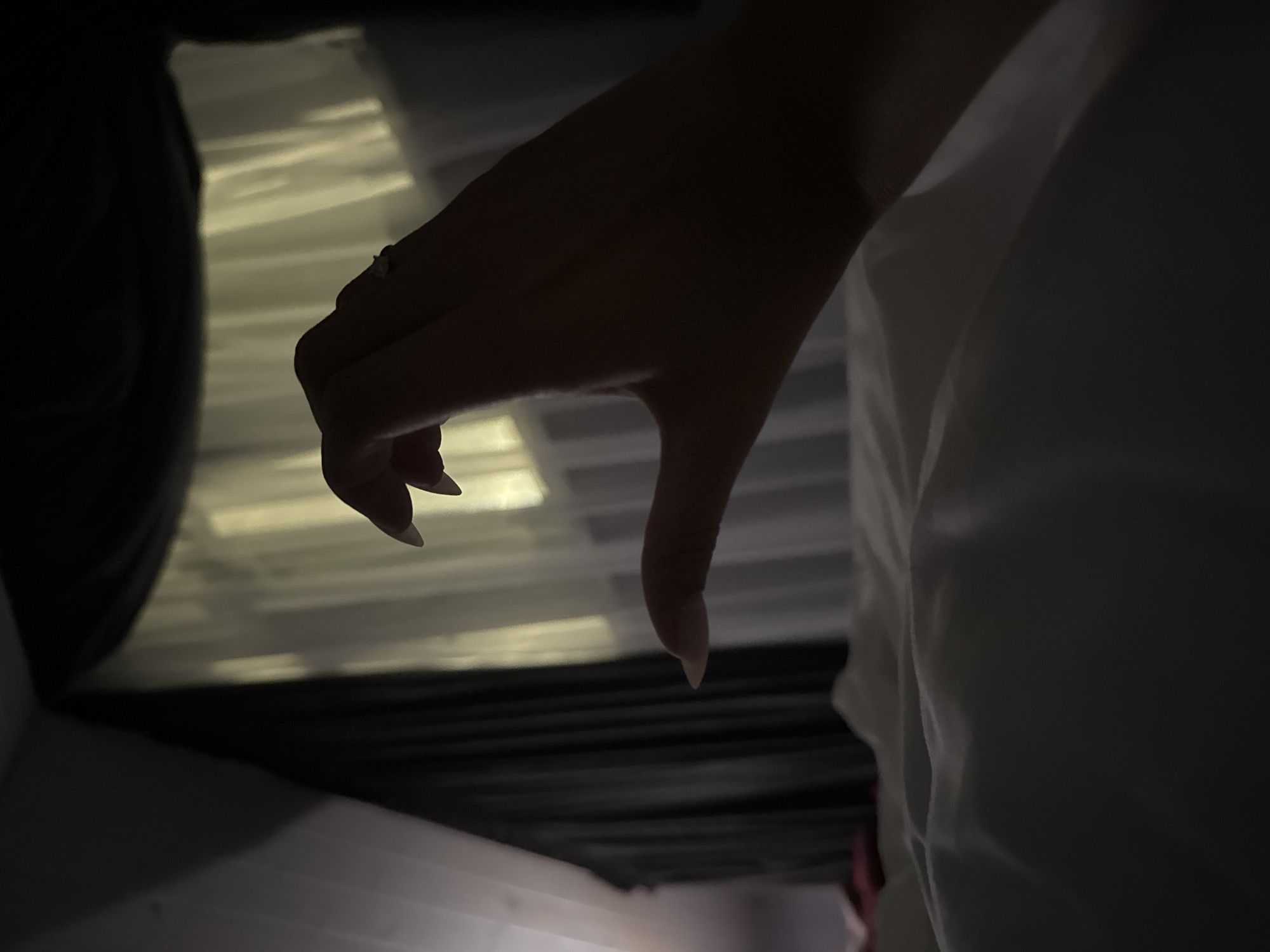 A close-up, low-light, over-the-shoulder shot shows a left hand forming a heart shape against a backdrop of sheer white curtains illuminated by a warm, yellow light. The hand, with long, manicured fingernails, is positioned in the foreground and casts a shadow on the white, rumpled sheets of a bed. Dark, heavy curtains frame the window, creating a somber atmosphere. The location is identified as Doma 1096 Km, Russia.