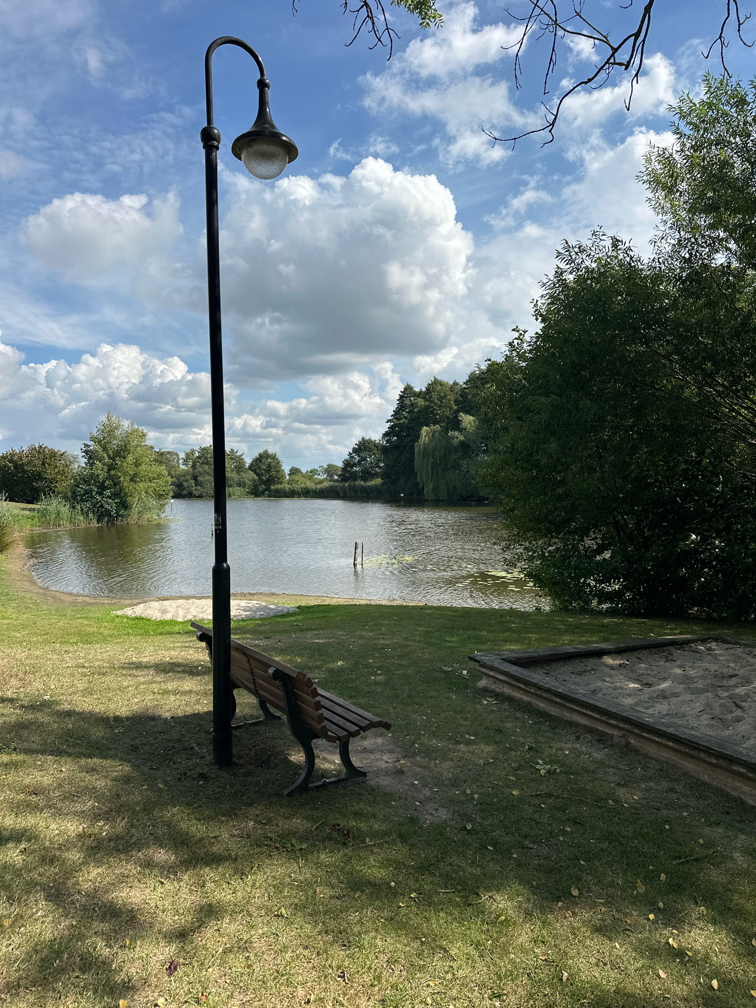 The image depicts a serene outdoor scene, likely in Samtgemeinde Thedinghausen, Germany, featuring a tranquil body of water fringed by lush green vegetation and trees. In the foreground, a wooden park bench is positioned on a grassy area, offering a place to rest. Adjacent to the bench is a sandpit, suggesting a recreational space. A tall, dark lamppost stands behind the bench, its curved arm supporting a vintage-style lamp. The sky above is a vibrant blue, dotted with fluffy white clouds, indicating pleasant weather. The water reflects the sky and surrounding greenery, adding to the peaceful ambiance. The overall impression is one of a well-maintained public park or recreational area, ideal for relaxation and outdoor activities.