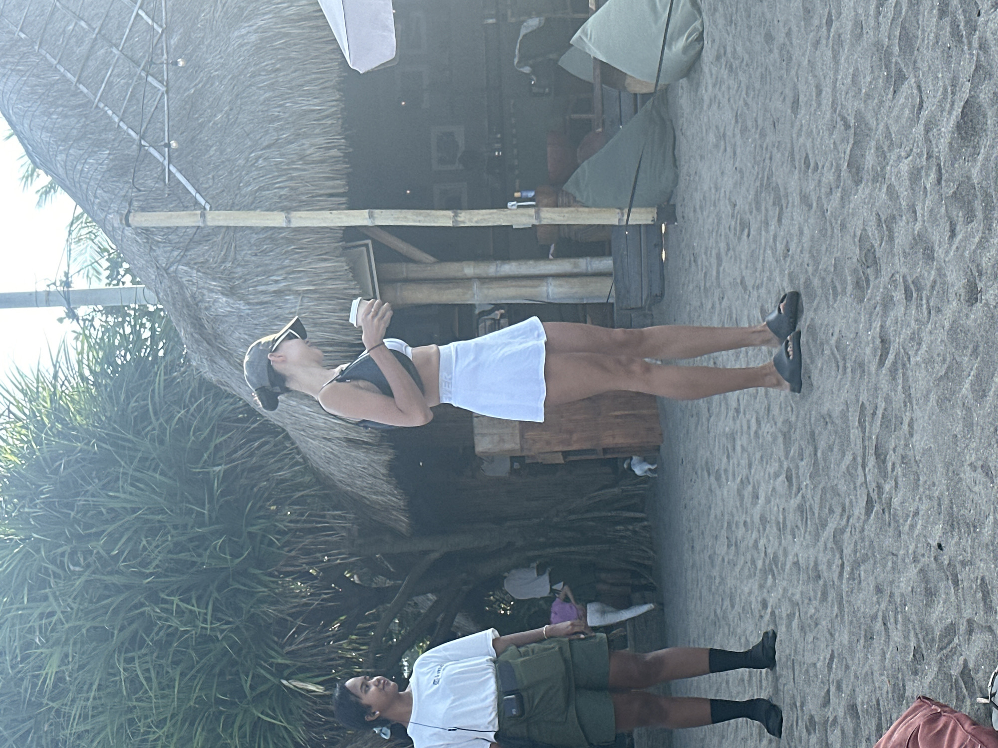 The image shows a beach in North Kuta, Indonesia, with two women standing on the sand. The woman on the left wears a white t-shirt, green shorts, and black socks and shoes. She has her hair tied back and is looking to her left. The woman on the right wears a white skirt and a dark top, and a dark baseball cap. She is holding a white cup and looking down. Behind them, there is a thatched-roof building with bamboo pillars, and beanbag chairs are visible on the sand. There are also trees and a clear sky in the background.