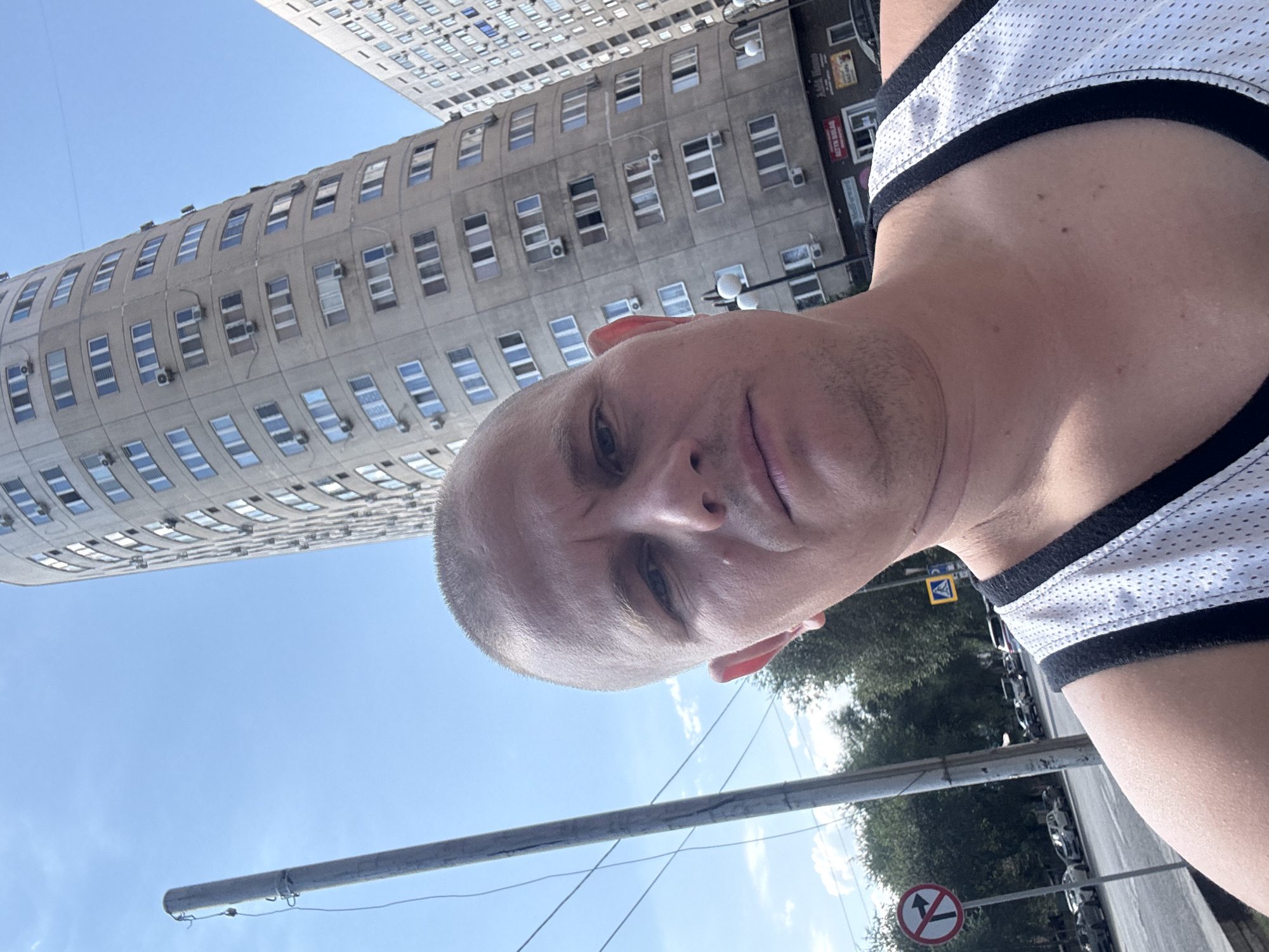 An outdoor, eye-level, close-up selfie shows a bald, light-skinned man with short facial hair in the foreground. He is wearing a white, sleeveless athletic shirt with black trim. Behind him, a tall, curved, grey apartment building with many windows and air conditioning units dominates the background. To the left, a utility pole with wires is visible, as is a traffic sign indicating no right turn. The scene appears to be a city street in Almaty, Kazakhstan, with trees and cars visible further in the background under a clear blue sky.