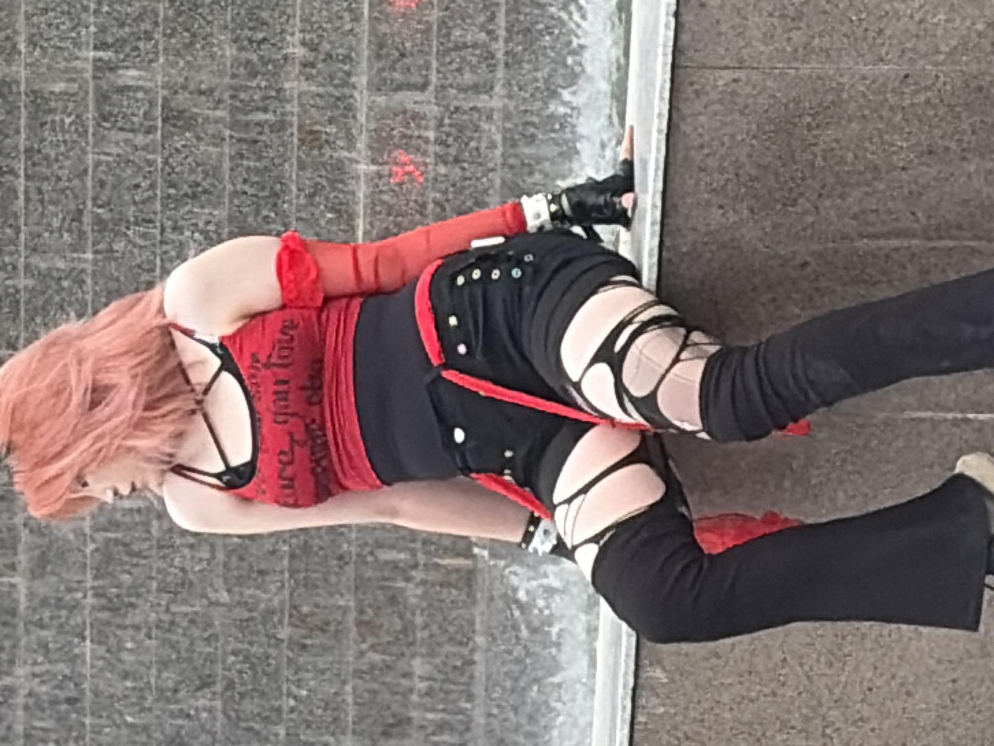 A person with bright pink hair, wearing a red tank top with a black bra and black shorts with ripped leggings, sits on a concrete ledge in front of a textured wall that appears to be a waterfall or a decorative textured surface. The person is also wearing black gloves and a spiked bracelet. The lighting is overcast. The location is Unknown, Unknown.