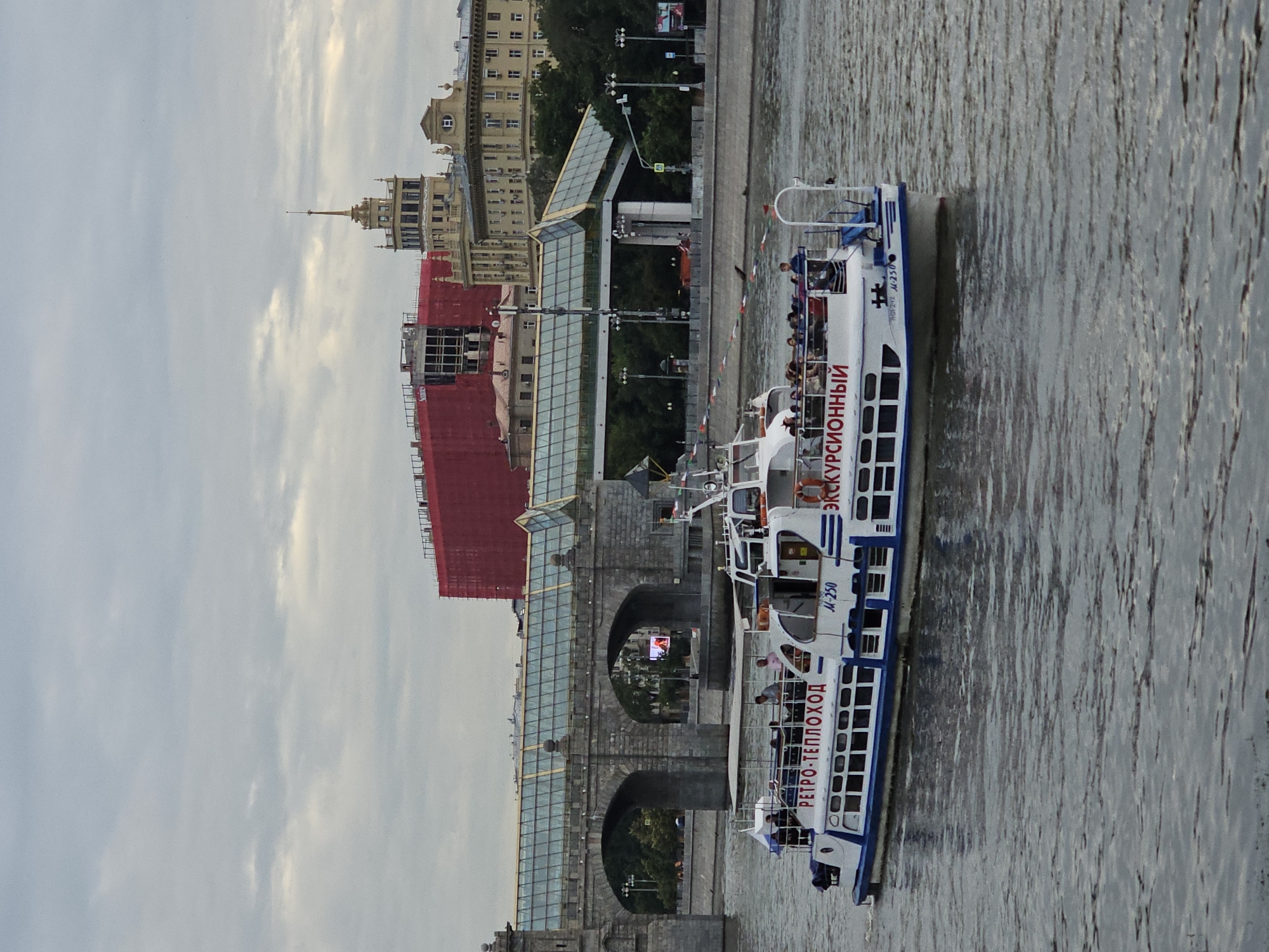 The image shows a tour boat on a river in Moscow, Russia, with a bridge and buildings in the background. The boat, named "Ретро-теплоход" (Retro-steamboat) and marked "M-250", is white with blue trim and has passengers on its upper deck. It is moving from left to right. In the background, a large, arched stone bridge with a modern glass walkway spans the river. Beyond the bridge, several buildings are visible, including a tall yellow building with a spire and a large building covered in red scaffolding. The sky is overcast. The river is wide and appears to be the Moskva River, given the presence of iconic Moscow architecture.