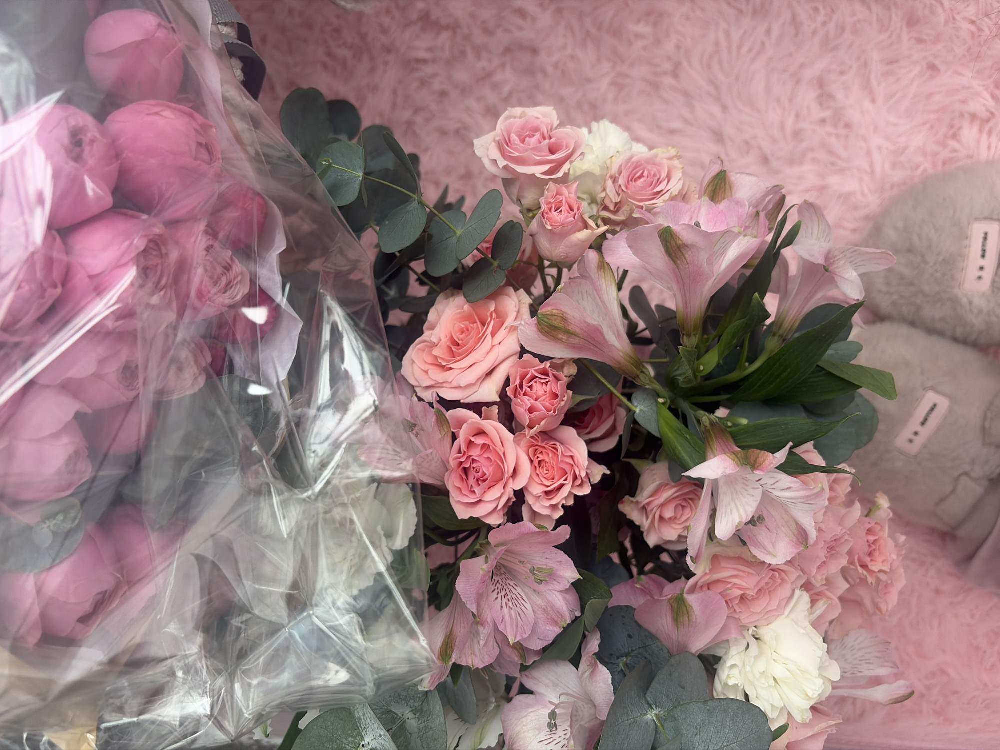 A close-up shot captures a bouquet of flowers, predominantly in shades of pink and white, arranged on a fluffy pink surface. The bouquet features roses, alstroemeria, and eucalyptus leaves. Part of the bouquet is enclosed in clear plastic wrap, revealing more pink flowers beneath. The scene appears to be an indoor setting. The image does not provide any information about the city of Unknown, Russia.