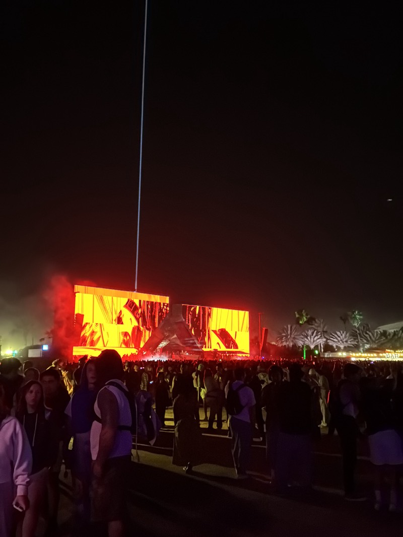 The image shows a large crowd of people at a music festival in Indio, United States, at night. A stage is lit with bright orange lights, displaying an abstract design. A tall, thin structure is visible behind the stage, reaching into the dark sky. Palm trees are visible on the right side of the image. The crowd is a mix of individuals wearing different clothing styles. There are visible traces of smoke or haze in the air near the stage.