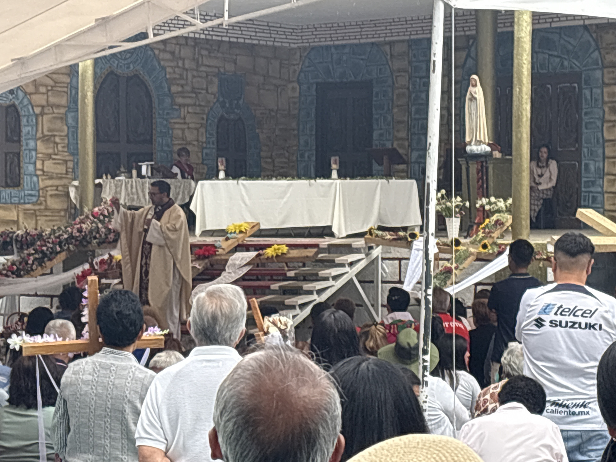 The image shows an outdoor Catholic Mass in what appears to be a public space. A priest in liturgical vestments stands on a flower-adorned stage delivering the service. Behind him is a large altar covered with a white cloth and what resembles a painted backdrop of a stone structure with archways. To the right of the altar is a statue of the Virgin Mary. Numerous people attend the Mass, their faces partially obscured as they are viewed from behind. Some are carrying wooden crosses decorated with flowers. A man in a white shirt with "Telcel Suzuki" printed on it is standing among the crowd. The location is likely Mexico City, Mexico, based on the presence of Telcel (a Mexican telecommunications company) branding and the general cultural context suggested by the religious event.