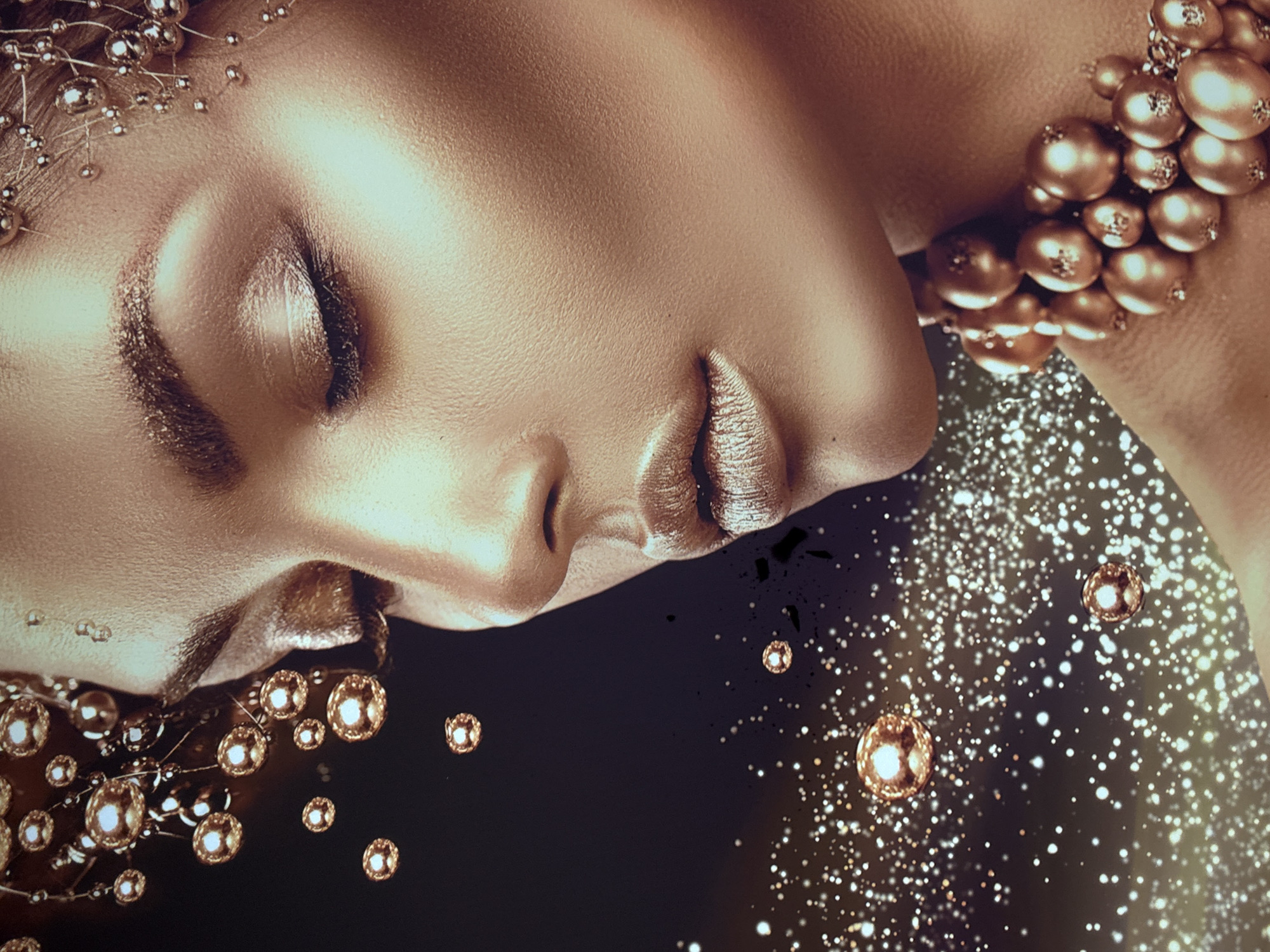 A close-up shot features a person with skin painted in a shimmering gold tone, in an unknown location. The person's eyes are closed and their head is tilted to the side, resting on a dark surface. The lips are also painted in a matching gold color. Adornments include beaded necklaces or accessories in the same gold hue, with some arranged around the head and others around the neck. The background is a dark color interspersed with small, golden spherical objects that resemble bokeh. Some dark areas are present that look like splatters or smudges.