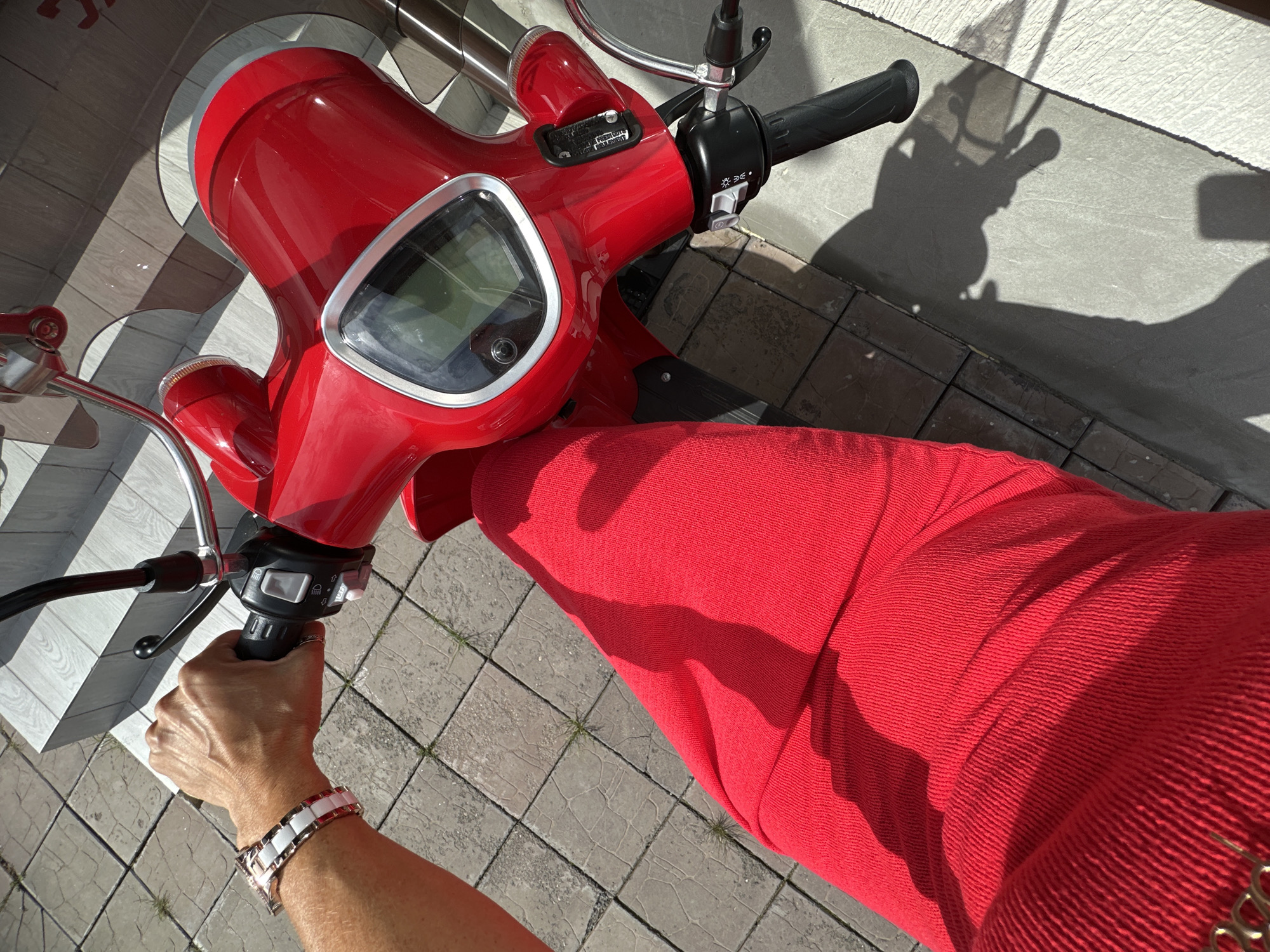 A first-person perspective shot captures a hand gripping the handlebars of a bright red scooter. The hand is adorned with a white and rose gold bracelet. The scooter's front panel, featuring a digital display, is prominent. A portion of the rider's red skirt is visible, draped over the scooter. The scene is set outdoors on a paved area composed of square tiles, with sunlight casting strong shadows. There is no indication of the city of Tyumen, Russia, or any other specific geographical location.