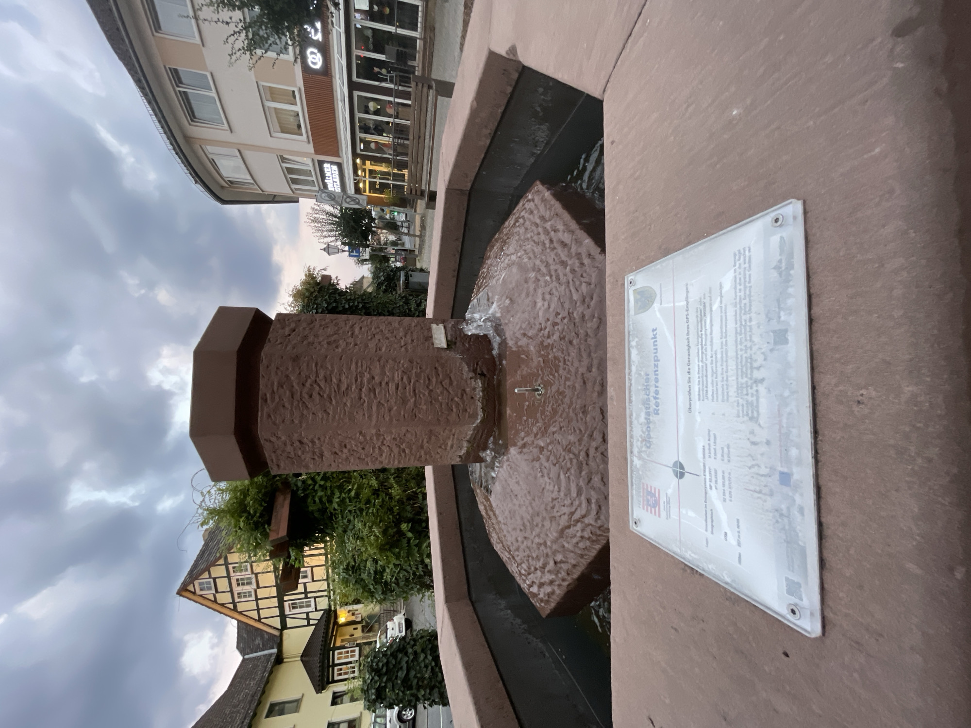 The image shows a public fountain in Neukirchen, Germany, identified by a sign on its base. The fountain is made of reddish-brown stone and features a tiered design with water flowing from the top. A sign in front of the fountain displays information in German, including coordinates and a compass rose, indicating it is a geodetic reference point. In the background, traditional German half-timbered houses are visible, along with a modern building with shopfronts and awnings. The sky is overcast, suggesting a cloudy day. There are no people or animals visible in the scene.