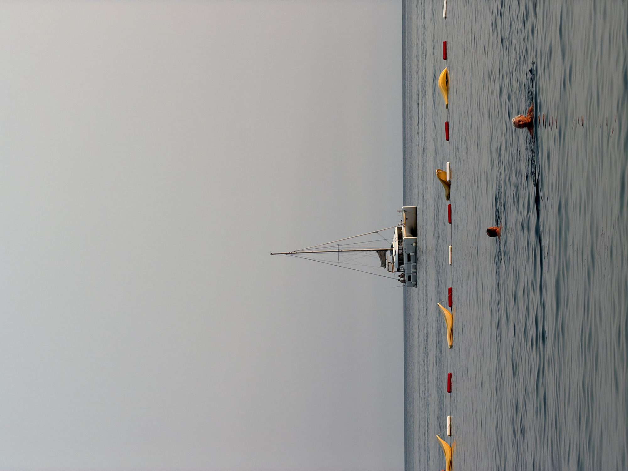 In the city of Prijevor, Montenegro, a calm, grey sea stretches to the horizon under a hazy, light grey sky. A white catamaran with two masts is anchored in the distance. Closer to shore, a line of buoys, alternating yellow and red, separates the swimming area from the open water. Several individuals are present in the water: two people are swimming in the foreground, their heads visible above the surface, and a few more figures are on the deck of the catamaran. The water is a deep, muted blue with gentle ripples.