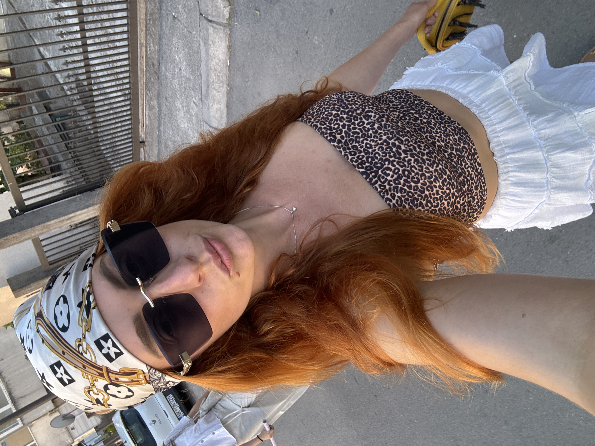 A woman with long, auburn hair stands outdoors, wearing a headscarf with a patterned design, dark sunglasses, a leopard-print bandeau top, and a white tiered skirt. She is holding a yellow bag in her right hand. In the background, a white car with a license plate is visible, as well as a metal fence and a sidewalk. The scene appears to be a street in Budva, Montenegro, during daylight.