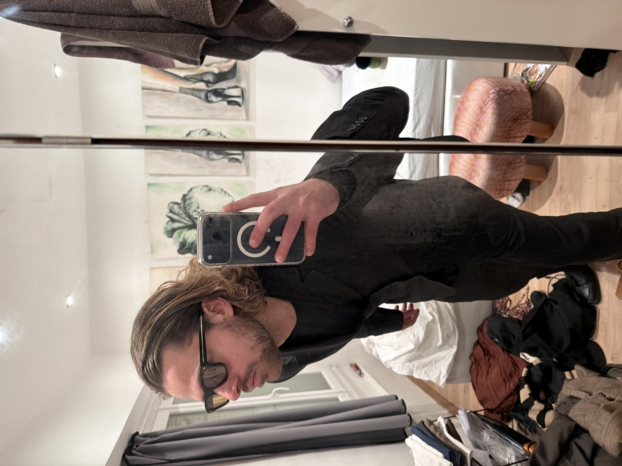 A male individual in his late 20s to early 30s with wavy, light brown hair and a beard is depicted in a mirror selfie taken indoors, located in a room within Paris, France. He is wearing dark sunglasses with gold accents and a dark suit jacket over a dark t-shirt. His right hand holds a smartphone, specifically an iPhone with a clear case featuring a white circular logo, angled to capture his reflection in a full-length mirror.
The background visible in the mirror's reflection includes a white wall with three framed artworks: one depicting a stylized head and two portraying high-heeled shoes. To the left, a window with closed dark grey curtains is visible. A white-sheeted bed is partially seen behind and to the right of the individual. The floor is light-colored wood, covered with various items of clothing in dark, brown, and striped patterns, alongside a small multi-tiered shelving unit stacked with folded garments. A small, pink, textured ottoman with light wooden legs rests on the floor to the right. A brown towel hangs on a fixture to the far right. The room is illuminated by overhead recessed lighting.