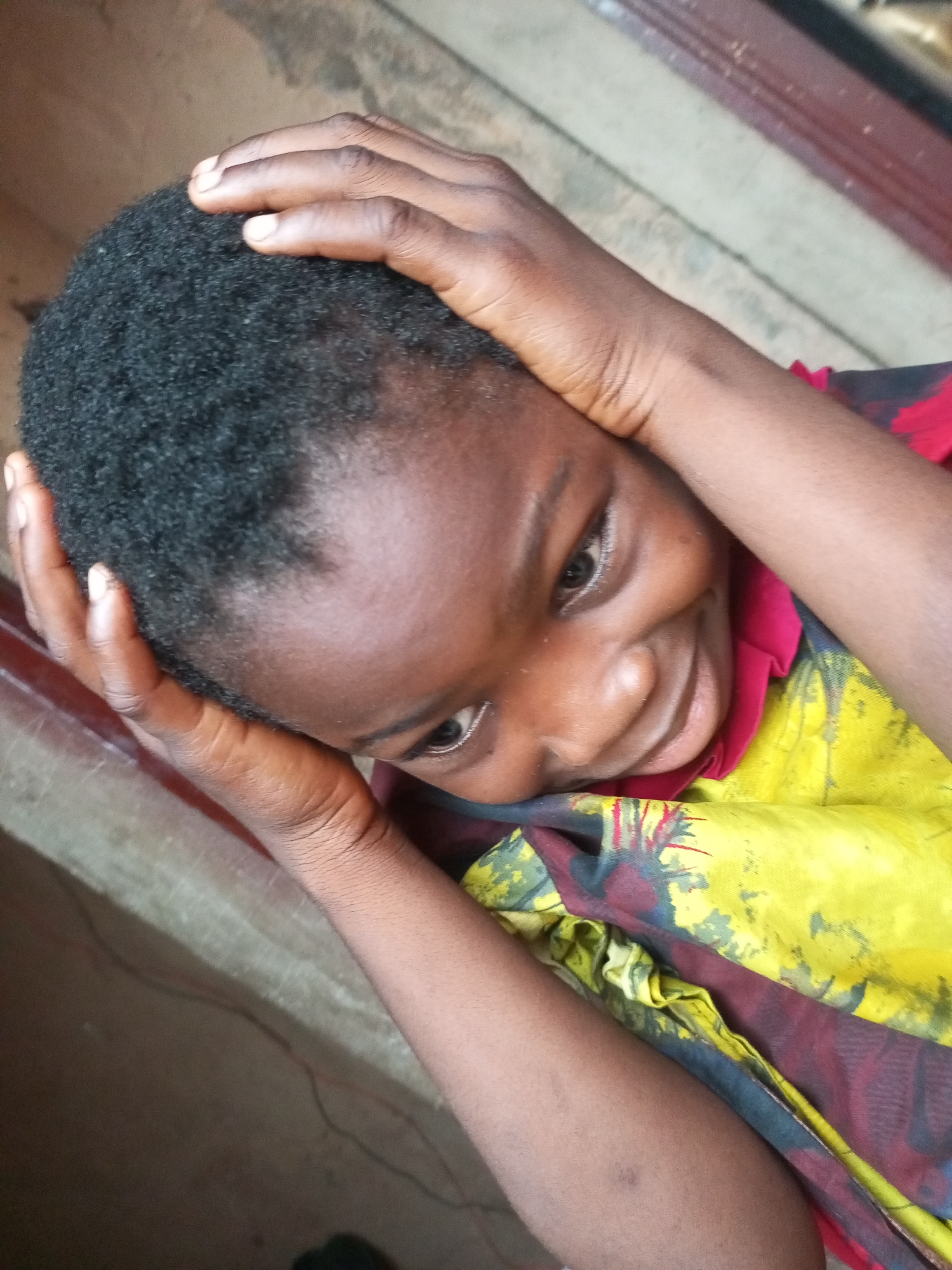 A close-up, high-angle shot shows a young Black person with short, dark, curly hair, resting their head on their hands. Their eyes are partially closed, and they have a slight smile on their face. They are wearing a yellow shirt with a dark, red, and blue pattern. The background appears to be a textured, light-colored surface, possibly concrete, with a dark, red wooden beam running diagonally across the upper right portion of the frame. The image is taken outdoors, with ambient lighting suggesting daytime. The scene is set in the city of Tempure, Nigeria.