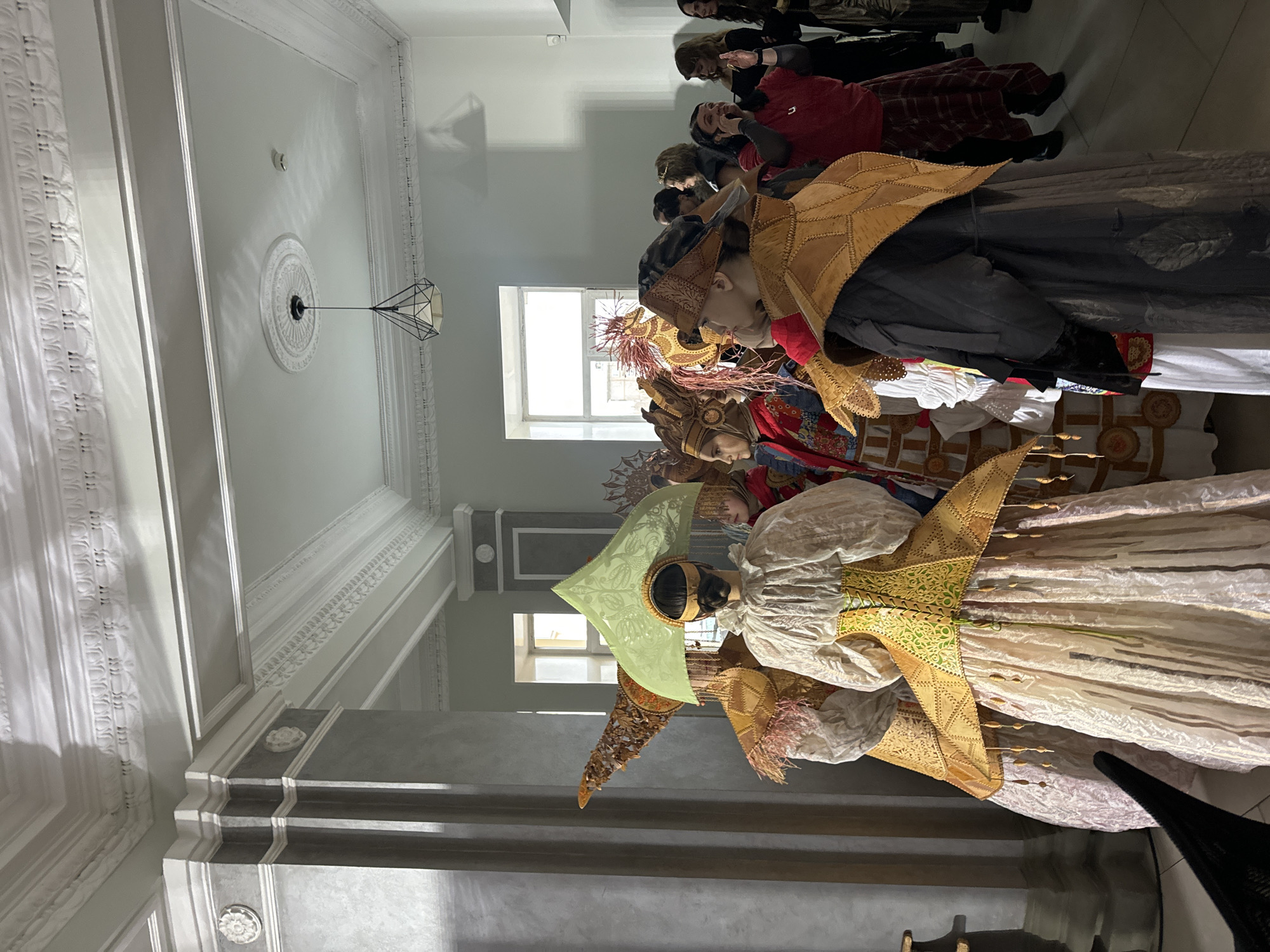 A group of people dressed in elaborate, stylized traditional costumes, featuring ornate headdresses and layered garments, are positioned in a long line. The location is indoors, and appears to be a museum or gallery setting based on the architectural elements such as decorative moldings on the walls, a patterned ceiling medallion, and large windows. The scene is brightly lit, possibly by natural light coming through the windows. The costumes are the primary subject of the image and they are the main focus. The location is in Krasnoyarsk, Russia.
