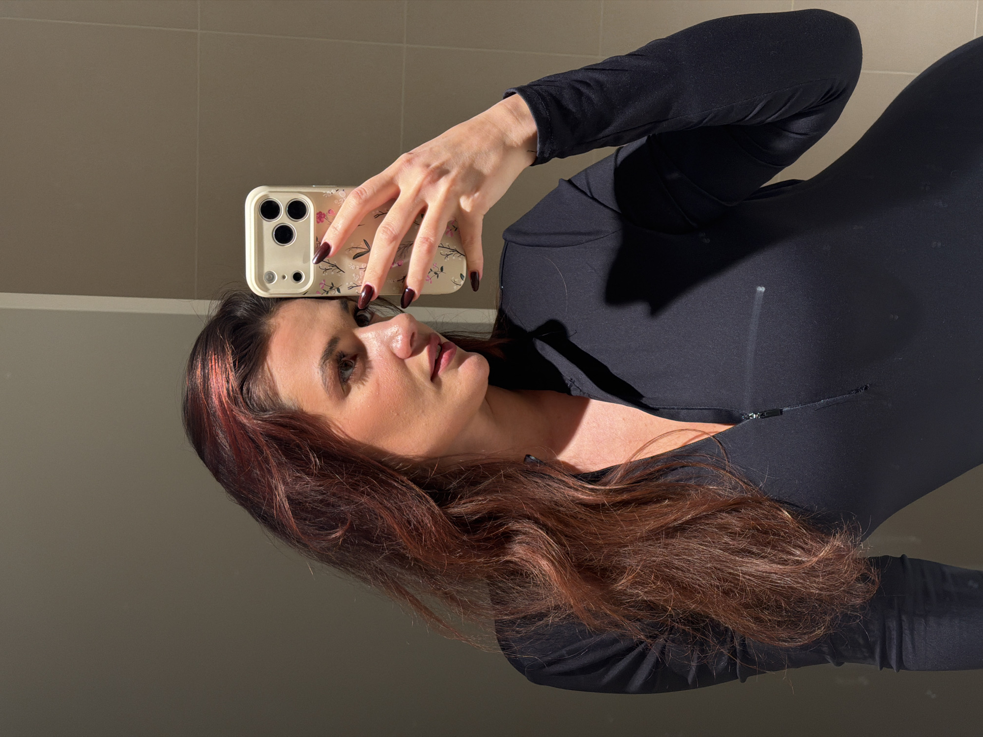 A female individual is depicted in a mirror selfie, captured from the upper torso, within an indoor environment featuring light brown tiled walls, likely located in Belgrade, Serbia. She has long, wavy reddish-brown hair with lighter highlights. The subject is wearing a dark, form-fitting, long-sleeved garment with a front zipper. Her right hand, featuring dark red manicured fingernails, holds a light-colored smartphone encased in a floral-patterned protective cover, positioned to photograph her reflection. A small tattoo is visible on her left wrist. Strong, directional lighting illuminates the right side of her face and hair, creating a bright highlight and casting shadows on the left side of her body and the background. The individual's gaze is directed slightly away from the camera.