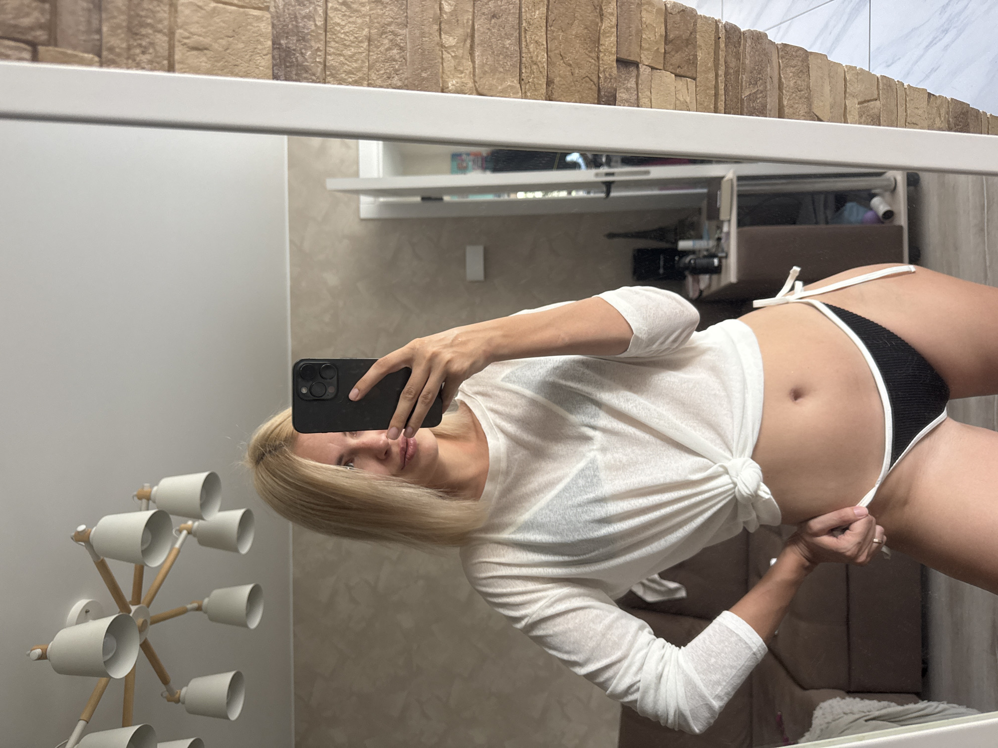 A woman with blonde hair takes a mirror selfie indoors. She is wearing a white long-sleeved shirt tied at the waist and black bikini bottoms. The background shows a sofa, a dresser with items on it, and a chandelier on the ceiling. The image does not provide information about the city of Saratov, Russia.