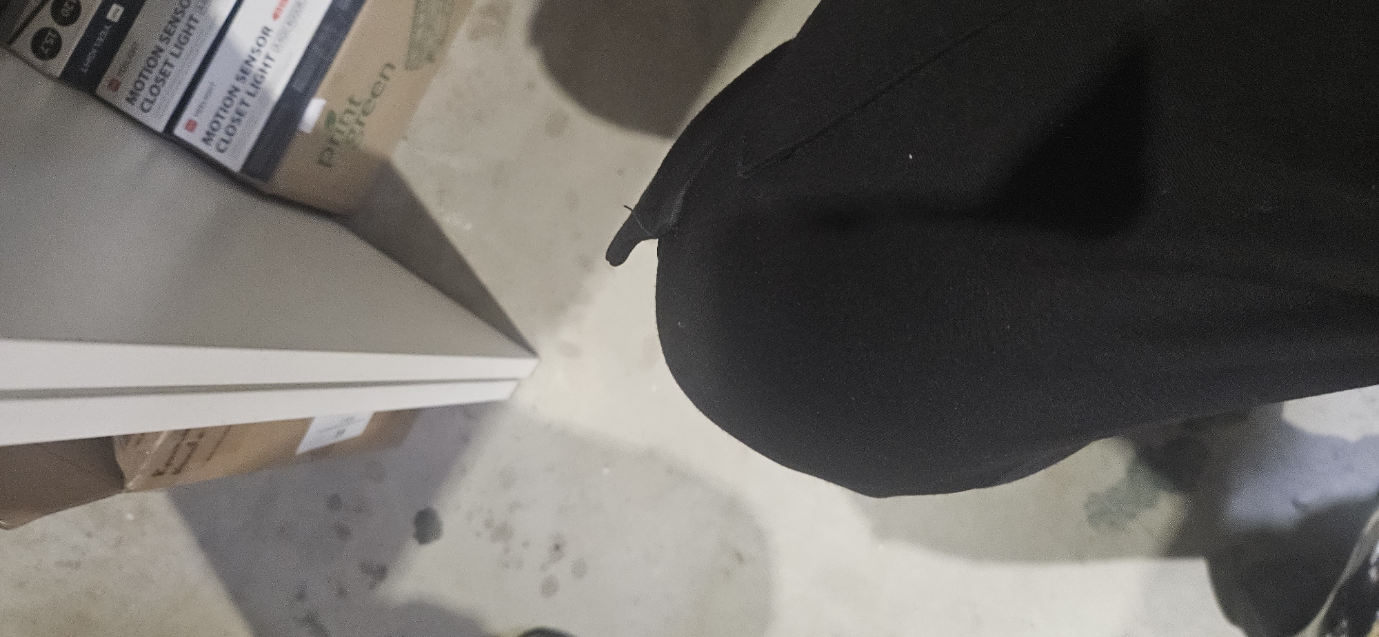 This image, taken in Plovdiv, Bulgaria, presents an indoor scene with a partial view of a person and several boxed items. On the right side of the frame, the lower body of a person, likely the thigh and upper leg, is visible, clad in black fabric, possibly trousers or leggings.
To the left, a light-colored, possibly white, flat surface or furniture item is prominent. Below and behind this, several cardboard boxes are stacked or placed on the floor. The most discernible text, repeated multiple times on one box, reads "MOTION SENSOR CLOSET LIGHT," suggesting the contents. Other visible text includes "PIR MOTION SENSOR CLOSET LIGHT," "TBEELIGHT," and "print green" on an adjacent box.
The floor is a light-colored, possibly concrete surface, marked with several dark stains or scuffs. The perspective is downward-looking, implying the person might be sitting, kneeling, or the camera is positioned low to the ground. No specific activity is clearly depicted, but the arrangement of boxes suggests an area for storage or organizing items. The lighting indicates an indoor setting.