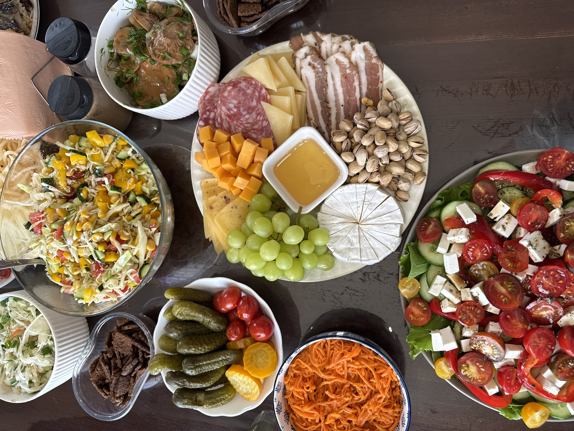 The image shows a table laden with an array of food items, likely a spread for a meal or gathering.
The central focus is a round platter featuring a selection of cheeses (including Brie, cheddar cubes, and sliced cheeses), salami, strips of bacon, pistachios, and a small dish of honey. Surrounding the platter are several bowls containing various salads and appetizers. These include:
* A salad with chopped cabbage, corn, cucumbers, tomatoes, and peppers.
* A bowl with shredded cabbage, green onions and fish.
* A bowl of marinated mushrooms.
* A bowl of pickled cucumbers and small tomatoes.
* A bowl with shredded carrots.
* A container with small croutons.
* A salad with tomatoes, feta, cucumbers, and red peppers.
Other items include table napkins and salt and pepper shakers.
The scene is indoors, set on a dark wooden table. There is no indication of people present.
The inclusion of marinated mushrooms, pickled vegetables, and shredded carrots is consistent with Russian cuisine. The city of Chaikovsky is in Russia, and these items are part of the types of food that are popular in this region.