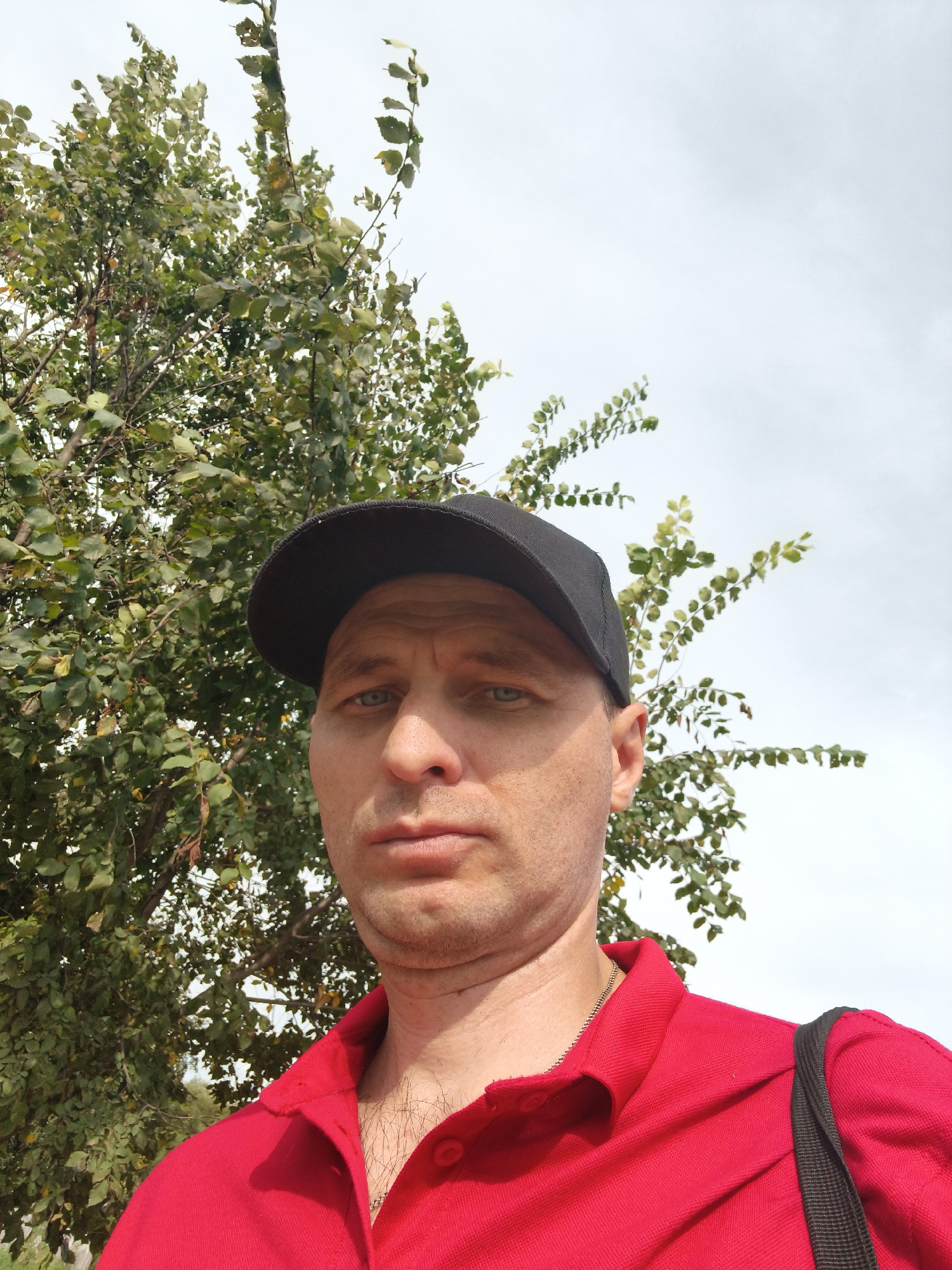 A man wearing a red collared shirt and a black baseball cap is pictured outdoors. He is looking towards the camera with a neutral expression. Behind him is a tree with green leaves and a light-colored, cloudy sky. The image appears to be a selfie taken in an outdoor setting. The location is stated to be in the city of Mokshan, Russia.