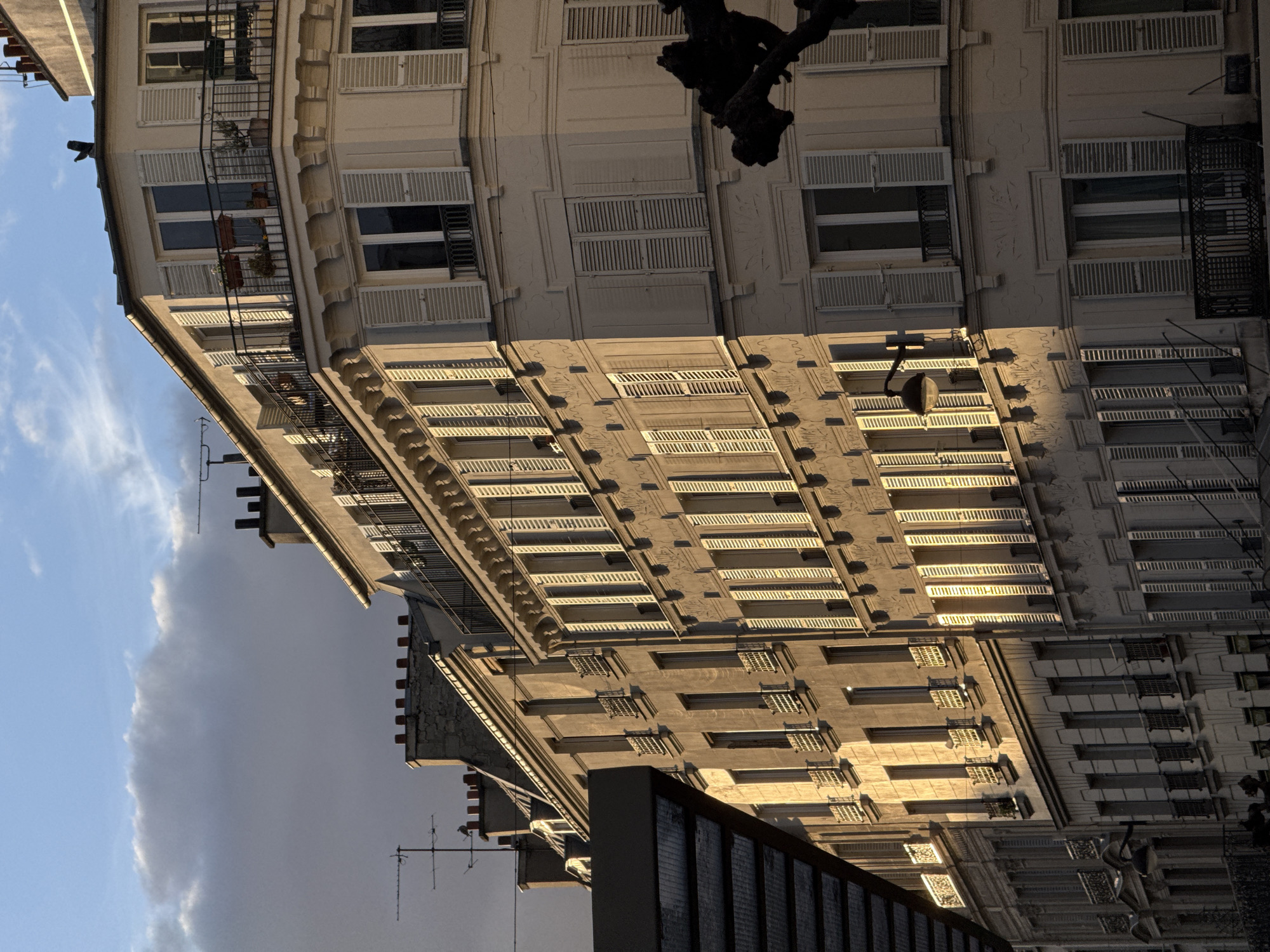 The image, when viewed upright, captures an urban street scene in Paris, France, dominated by several multi-story buildings under a sky with mixed cloud conditions. The central and most prominent structure is a large, light-colored building, characteristic of Haussmannian architecture, with multiple floors of windows. Most windows are fitted with white louvered shutters (jalousies), some closed and others open. Several floors feature ornate black wrought-iron balconies, some adorned with potted plants.
A warm, golden sunlight illuminates the right-facing facade of this main building, creating long, distinct shadows from the balconies, window recesses, and architectural details. This lighting suggests either late afternoon or early morning. The left side of the building and other structures in the background are cast in shadow. Above the buildings, the sky transitions from bright blue with wispy white clouds on the right to a larger, darker grey cloud mass towards the upper left. Several antennas are visible atop various rooftops. A dark, gnarled tree branch extends into the upper right of the frame, partially obscuring the main building.
In the lower foreground, the upper section of a darker, more modern-looking building with a gridded facade is visible. Further in the background, another traditional building with smaller, recessed windows is also present, partly touched by the sunlight. No people or specific activities are depicted, contributing to a serene, static composition. A faint, illegible street sign can be observed on one of the buildings.