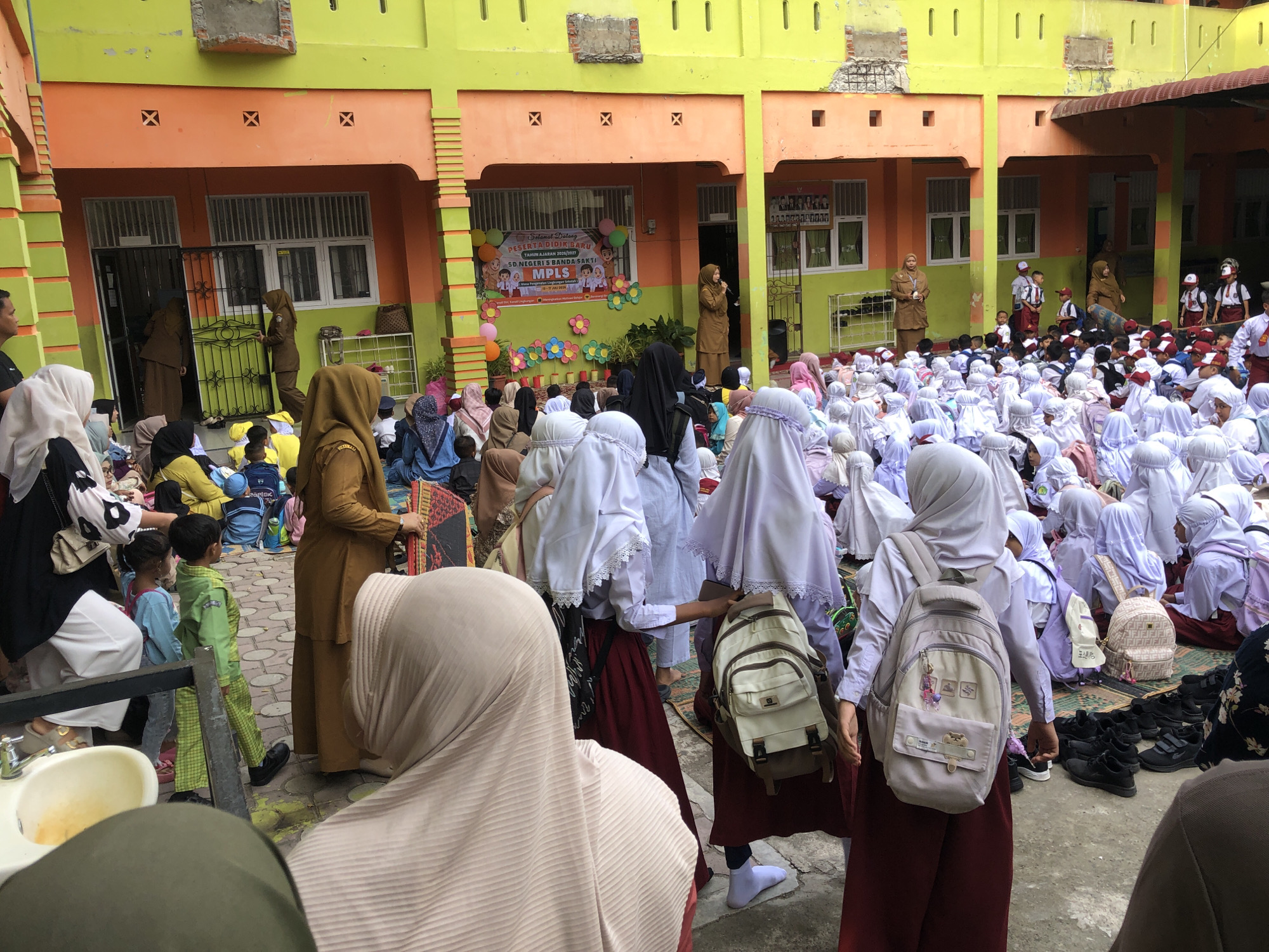 The image depicts a courtyard or outdoor gathering area of a school in Lhokseumawe, Indonesia. The building has a vibrant yellow upper level and an orange lower level with green accents. There are several individuals present, primarily students dressed in school uniforms, many of whom are wearing headscarves (hijabs). Some adults, presumably teachers or parents, are also visible.
In the foreground, a child in a green uniform is walking, looking to the right. Behind them, a woman in a tan hijab and matching outfit is standing. A group of children are seated on the ground in the middle ground, facing towards a building with a banner that reads "Selamat Datang Peserta Didik Baru" (Welcome New Students). Another banner appears to advertise "SD Negeri 5 Banda Sakti" (State Elementary School 5 Banda Sakti).
The ground is paved with patterned tiles. Various bags, backpacks, and shoes are scattered around the area. The overall atmosphere suggests an organized event, likely the beginning of the school year, with students and adults gathered for an orientation or welcome ceremony. The presence of a microphone suggests a speech or presentation is taking place or has just concluded.