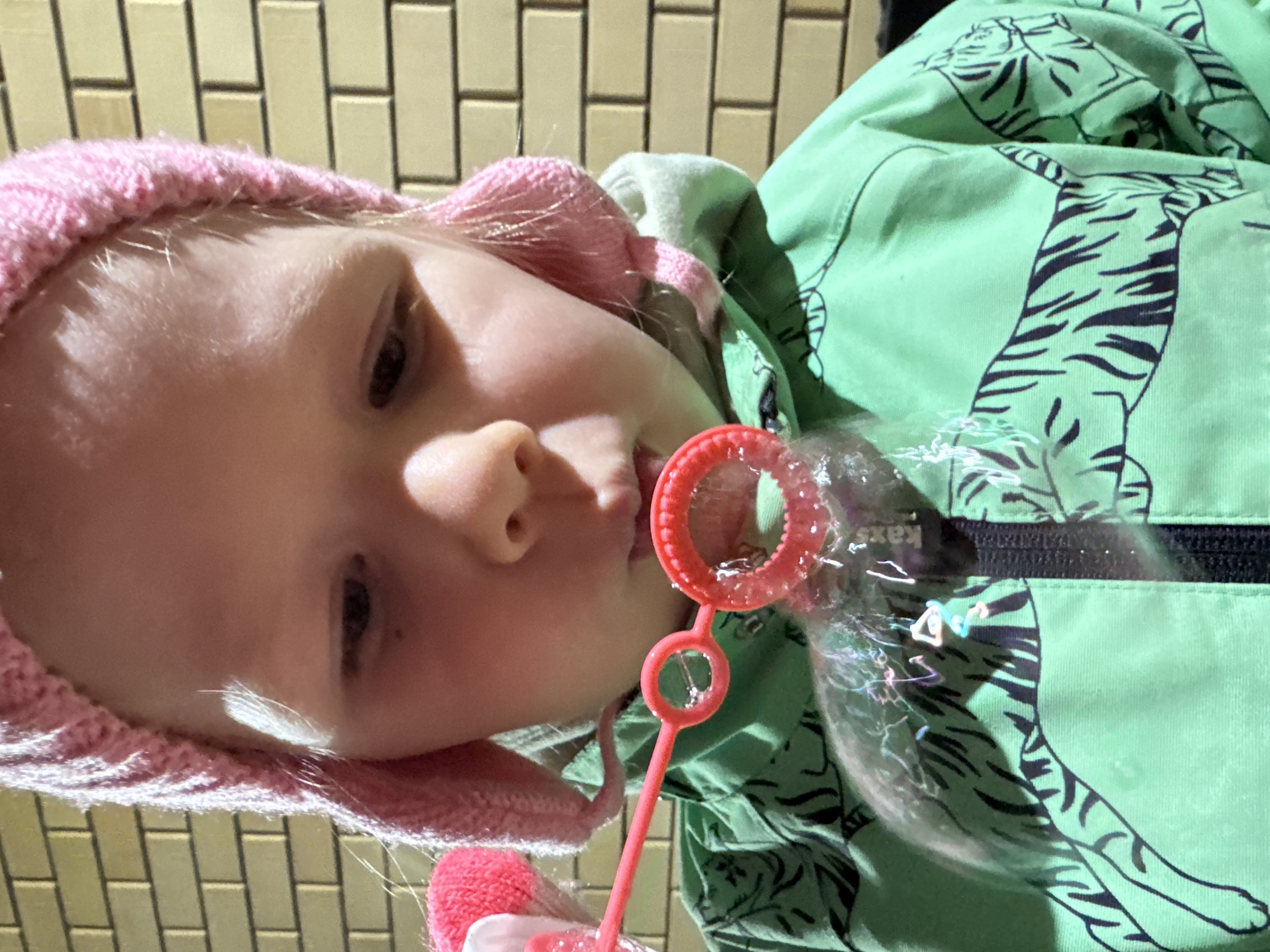 A young child is blowing bubbles outdoors. The child is wearing a pink knit hat and a green hooded jacket with black tiger stripe designs. A red bubble wand is positioned in front of the child's mouth, with a bubble partially formed. The location is Ферма, Russia, discernible from the "kaxs" logo present on the jacket. The background appears to be a wall or structure with a rectangular tile pattern.