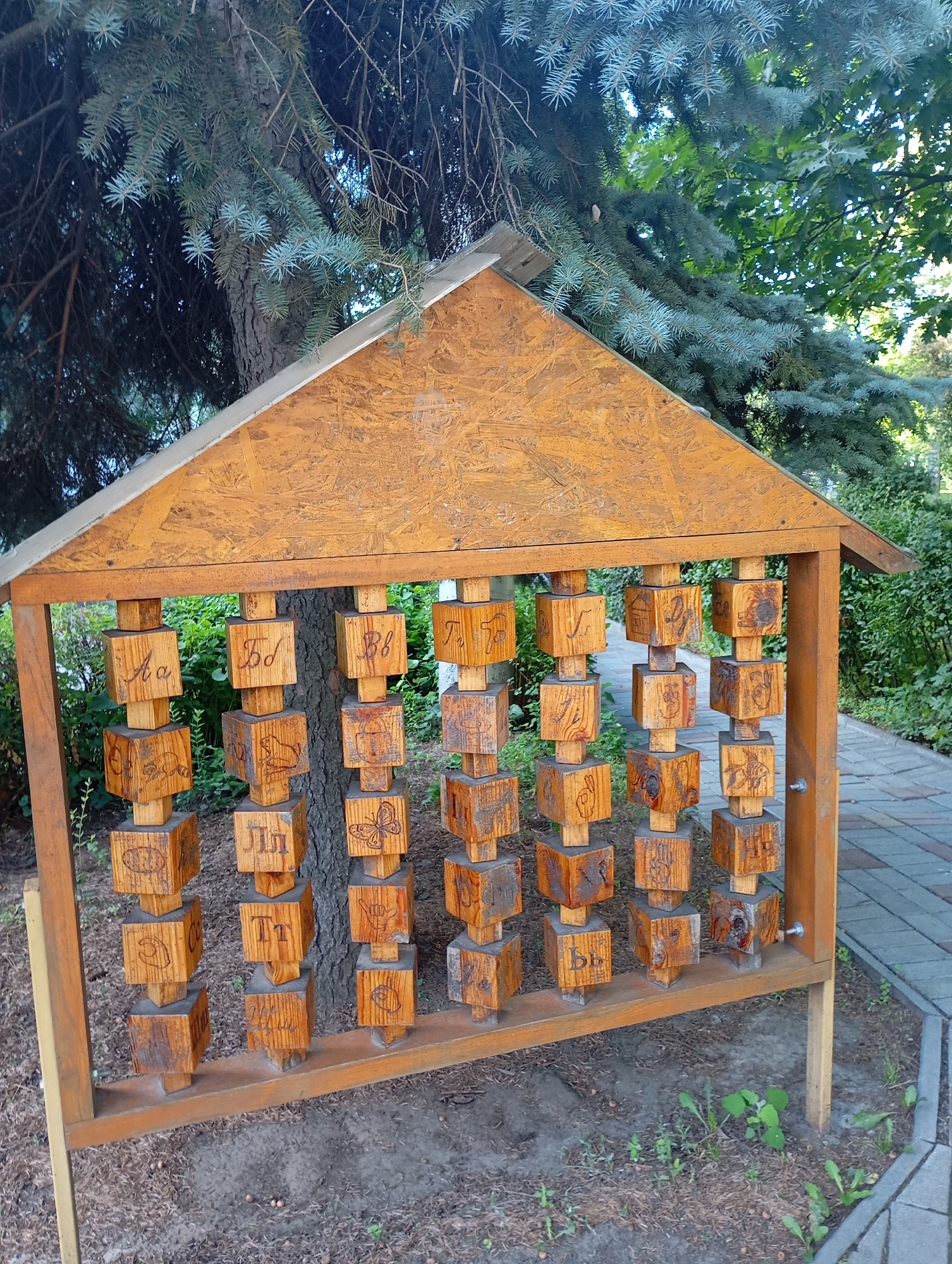 An outdoor wooden alphabet structure with carved wooden cubes featuring Cyrillic letters and images is situated in a park-like setting in Kharkiv, Ukraine. The structure is triangular in shape, resembling a small house with a roof made of oriented strand board. Suspended vertically from the roof are several rows of wooden cubes, each carved with a letter and a corresponding image. For example, one cube shows the Cyrillic letter "Aa" paired with a drawing of a bird, and another shows "Bb" with a drawing of a cup. The structure is supported by wooden posts and is surrounded by greenery, including a large conifer tree and bushes. A paved walkway runs alongside the structure. The ground is covered with dry leaves and some sparse vegetation. The scene is well-lit, suggesting it is daytime.