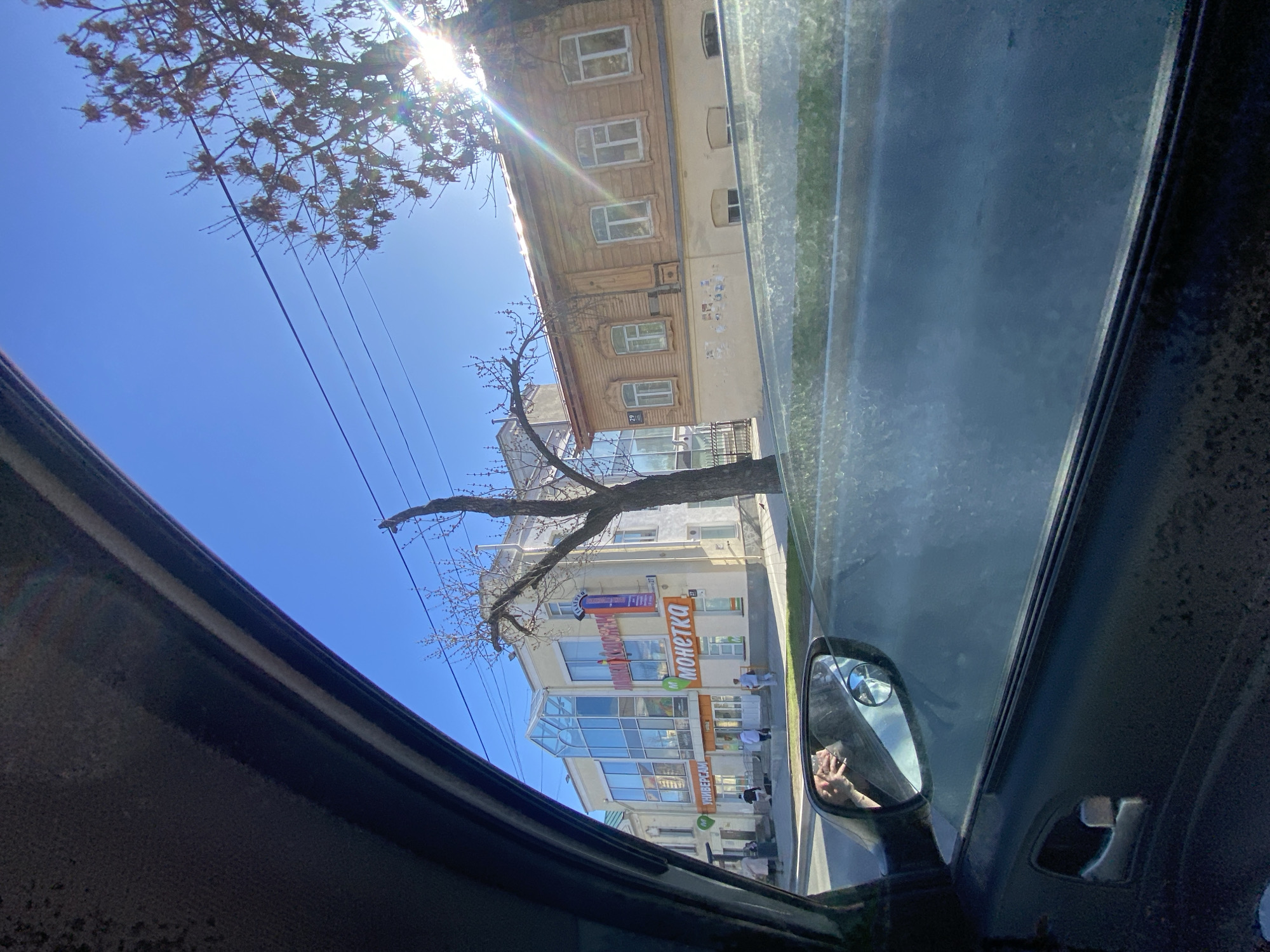 View from inside a car in Yekaterinburg, Russia. Outside the car window, there is a street with buildings on both sides. One building has a sign with "MOHETKA" written on it. There are a few people walking on the sidewalk. Power lines and a tree are visible against a bright blue sky. The sun is shining through the branches of a tree. The car's side mirror reflects a partial view of the driver's hand holding a phone.