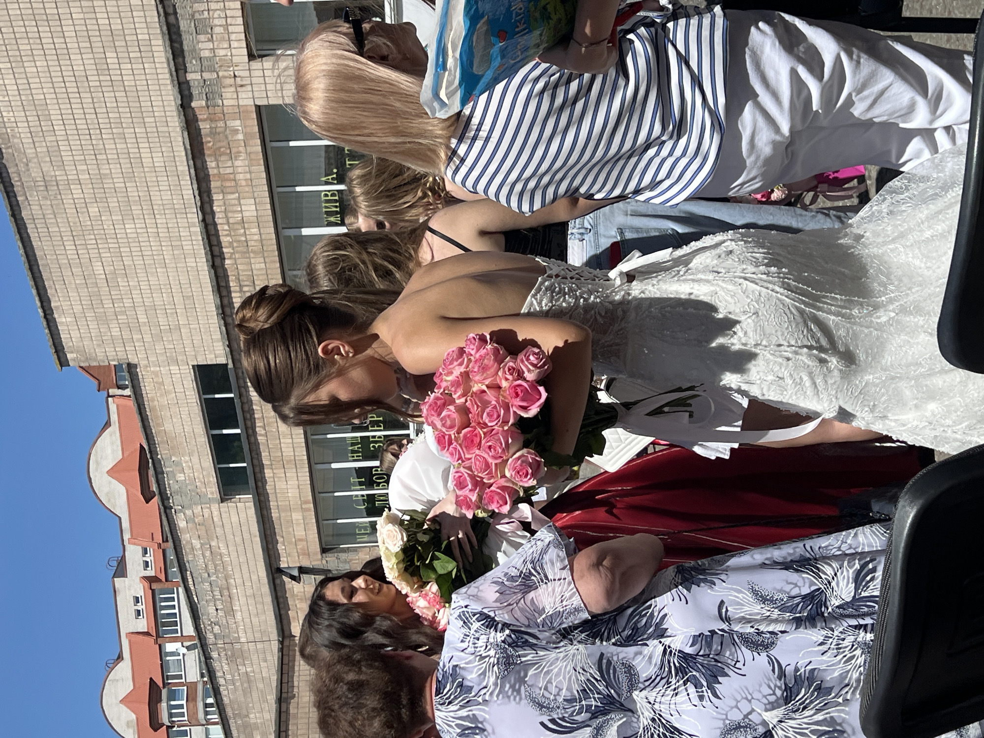 This is a vertical, full-body shot of a group of people gathered outdoors, likely for a celebration. The central subject is a young woman in a white, sleeveless dress, holding a large bouquet of pink roses. She is leaning forward, her face partially obscured as she receives or admires the flowers. Her hair is pulled back in a bun.
To her left, a woman with blonde hair is wearing a blue and white striped shirt and white pants. She is holding a colorful bag. Behind the woman in white, other individuals are visible, including one person in a red garment and another wearing a patterned shirt.
The background features a brick building with multiple windows. Some of the windows have signs with text. One sign reads in Cyrillic, "ЖИВ. А. Т." and another appears to say, "НЕН СВІТ НАЦ. ЛЮБОВ ЗВЕР." The building has a light-colored facade and a red-tiled roof with decorative architectural elements. The sky above is clear and blue, suggesting a sunny day. The overall atmosphere seems festive and joyous, possibly a wedding or graduation event, given the formal attire and the abundance of flowers. The image is taken from a slightly elevated perspective.