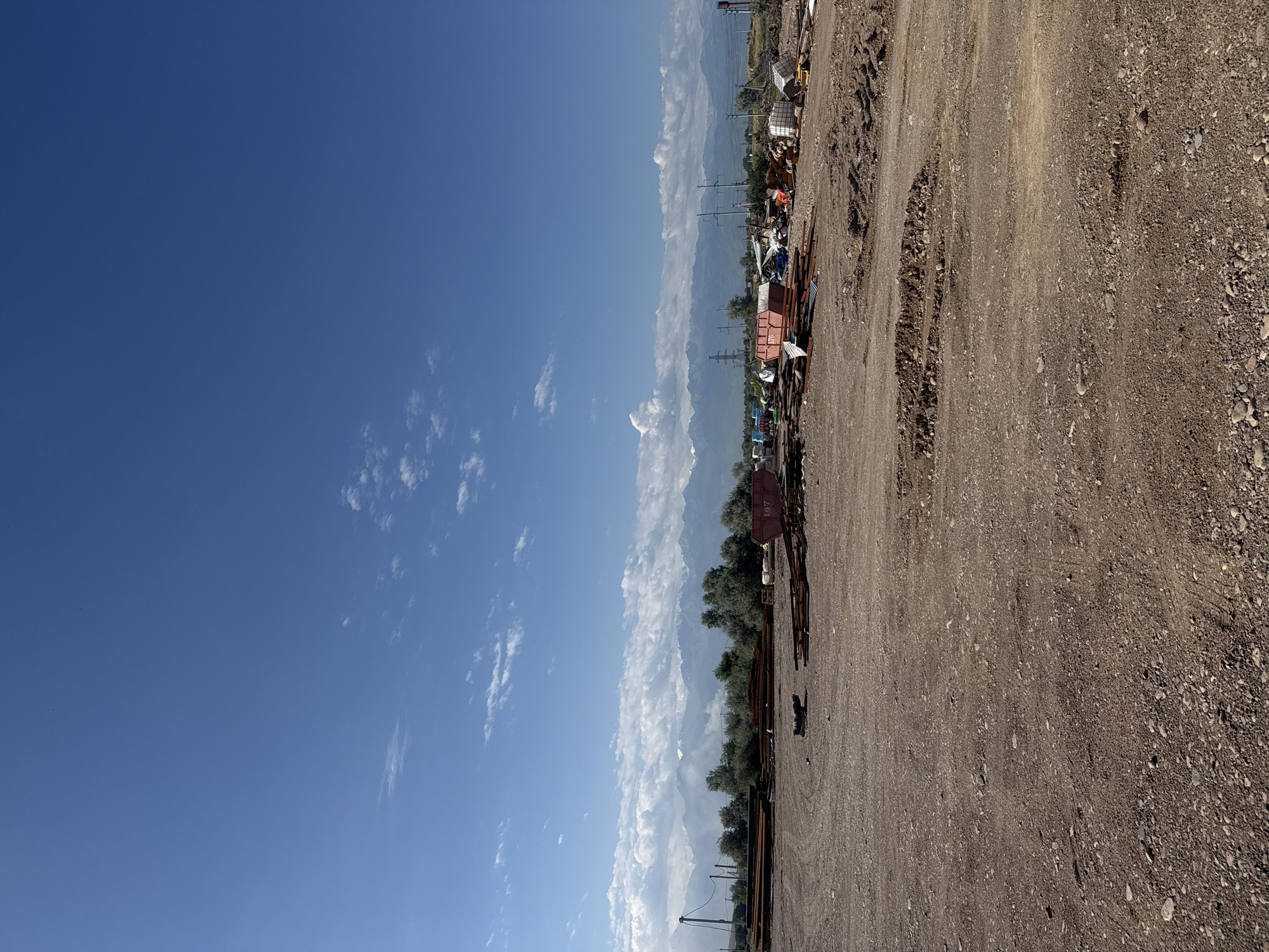 The image shows a desolate outdoor industrial area under a clear blue sky with scattered white clouds. In the foreground, a rough, dusty, and rocky ground is visible. In the midground, there are piles of rusted metal pipes and beams, along with various containers and equipment, suggesting a scrap yard or construction site. Some of the containers are red, and one has the number "197" visible. Behind the industrial debris, a line of trees separates the foreground from a distant mountain range. The mountains are rugged and appear to have snow-capped peaks. Power lines and transmission towers are also present in the middle ground and background, indicating the proximity of infrastructure. The scene is devoid of people and any signs of recent activity, conveying a sense of abandonment or inactivity within an industrial zone on the outskirts of Köközek, Kazakhstan.