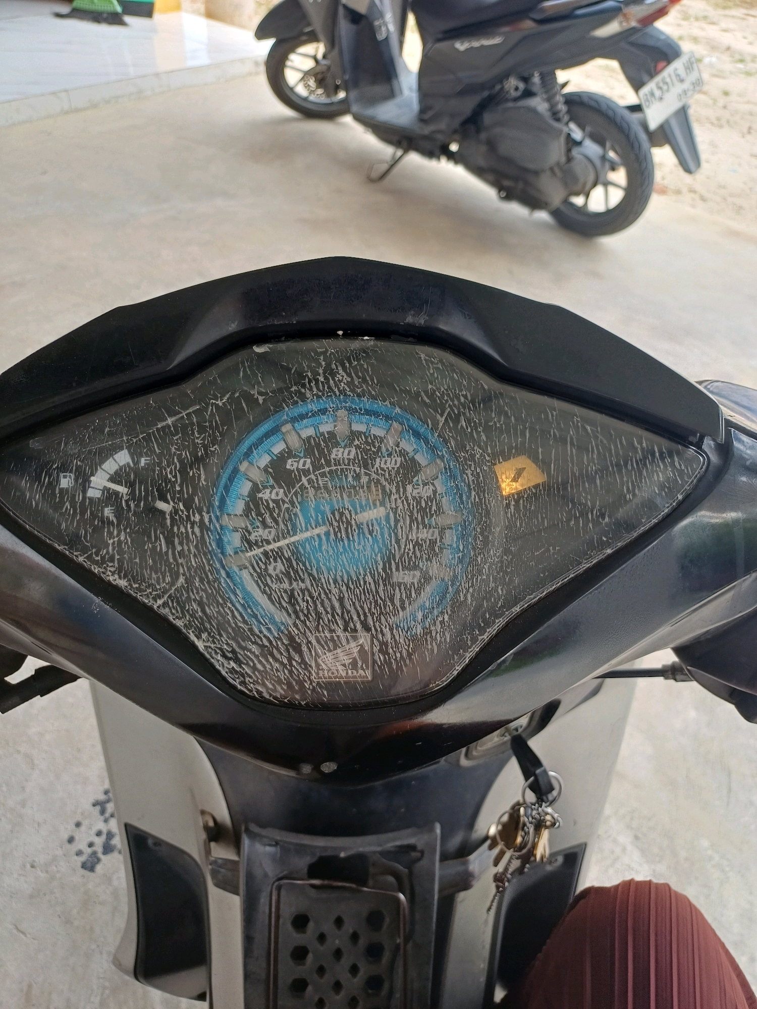 The image shows a close-up view of a cracked motorcycle dashboard, with a speedometer indicating a speed of approximately 40 km/h and a yellow warning light illuminated. In the background, another motorcycle is parked. The scene appears to be outdoors, possibly in a residential area or a parking lot, with a tiled floor and a sandy ground visible. The context of Dumai City, Indonesia, is provided by the user.