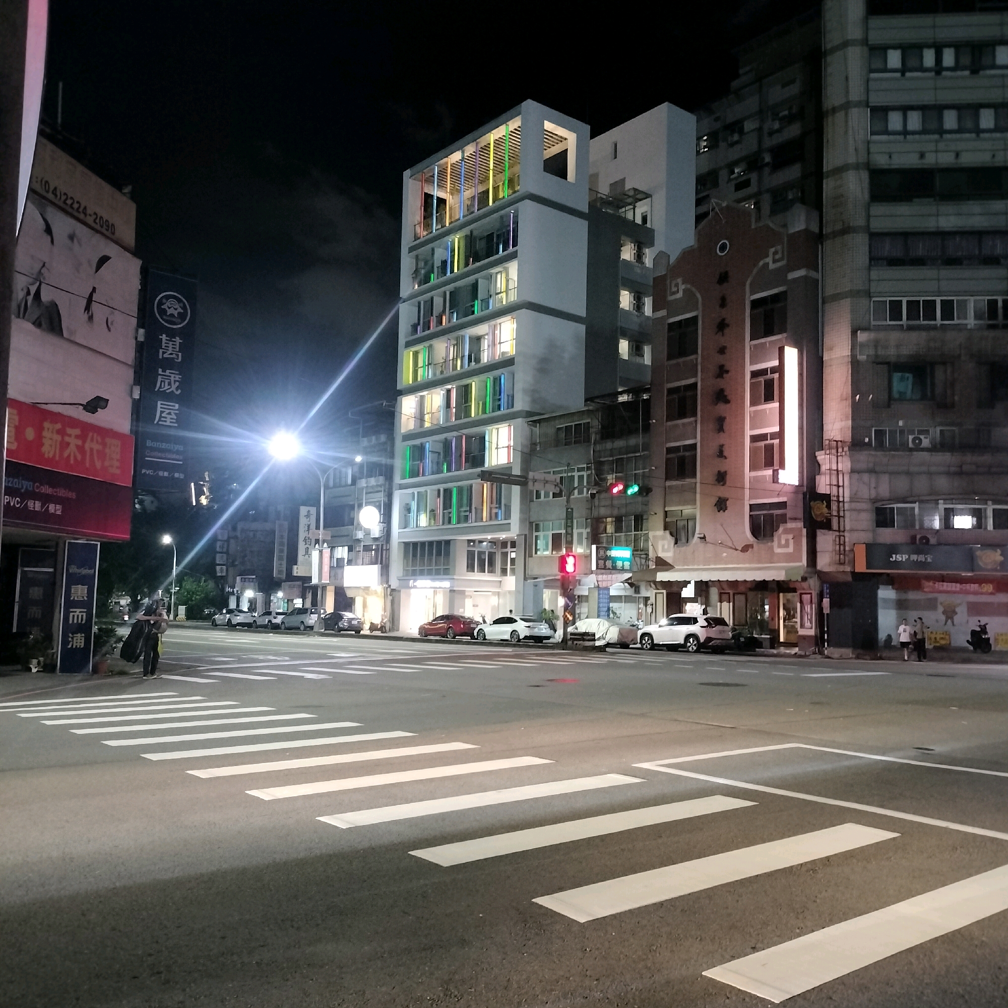 A night scene captures an intersection in Taichung, Taiwan, featuring a modern, multi-story building with colorful illuminated windows contrasting with older, adjacent buildings. The street is paved with asphalt and marked with white crosswalks. Several cars are parked along the street or are in motion, including a white sedan, a red sedan, and a white SUV. Pedestrians are present; one person stands near the curb holding a large black trash bag, and two others walk on the sidewalk further down the street. Streetlights illuminate the scene, casting light rays across the road and buildings. Signs with Chinese characters are visible on the buildings, indicating businesses. The sky is dark, suggesting it is late evening or night.