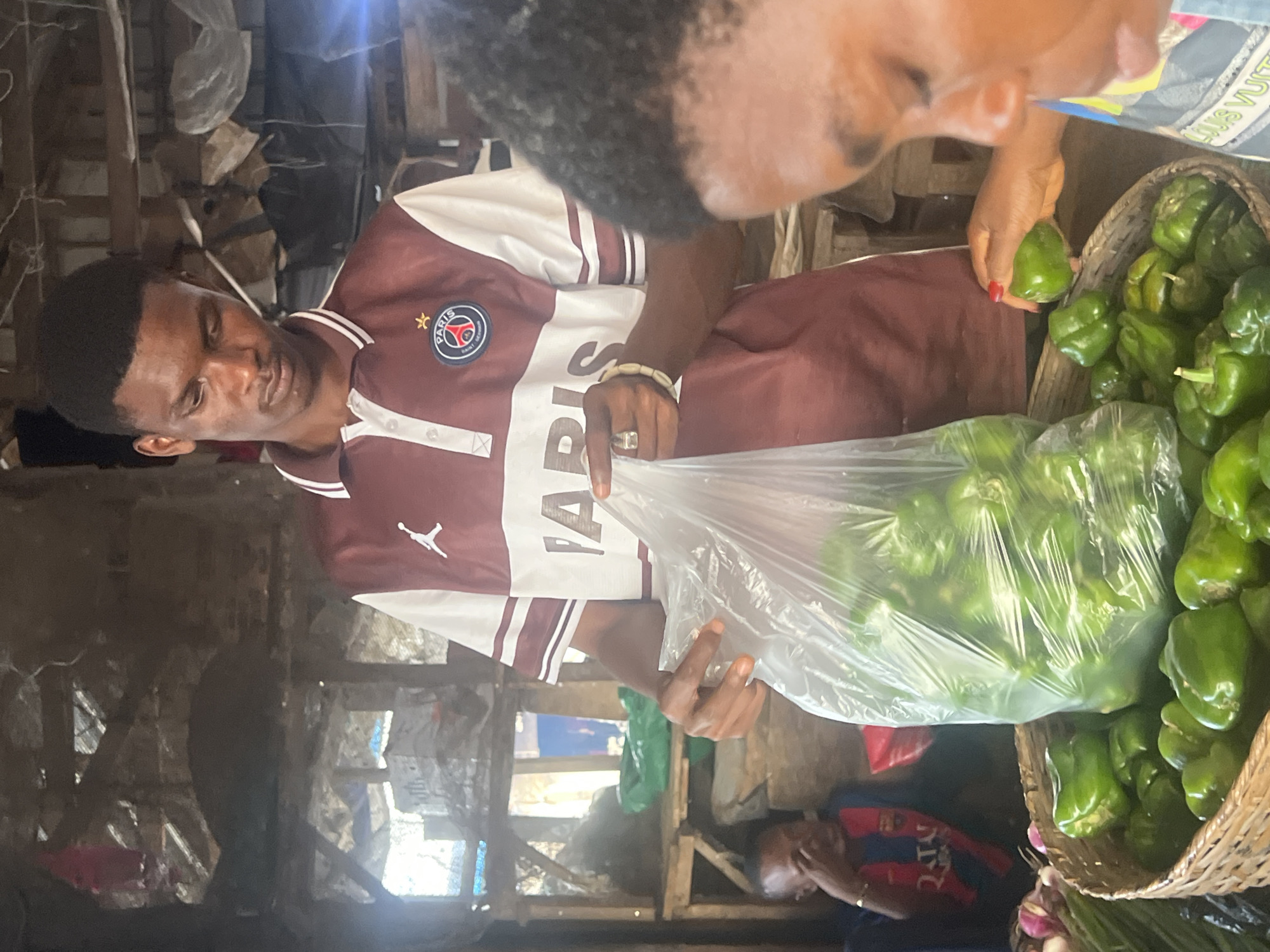 In the city of Oko Amakom, Nigeria, a young man stands at a market stall, holding a clear plastic bag filled with green bell peppers. He is wearing a maroon and white polo shirt with a Paris Saint-Germain logo and an Air Jordan logo. His right hand, adorned with a ring and a beaded bracelet, is extended, pointing towards the peppers. In the foreground, a basket overflows with more green bell peppers. To the right, a woman is partially visible, her hand reaching for a pepper from the basket. In the background, another person is seated, appearing to be looking at their hand. The stall is constructed of rough wood and metal, with shelves holding various items, some covered with dark cloth. The overall environment suggests a bustling local market.