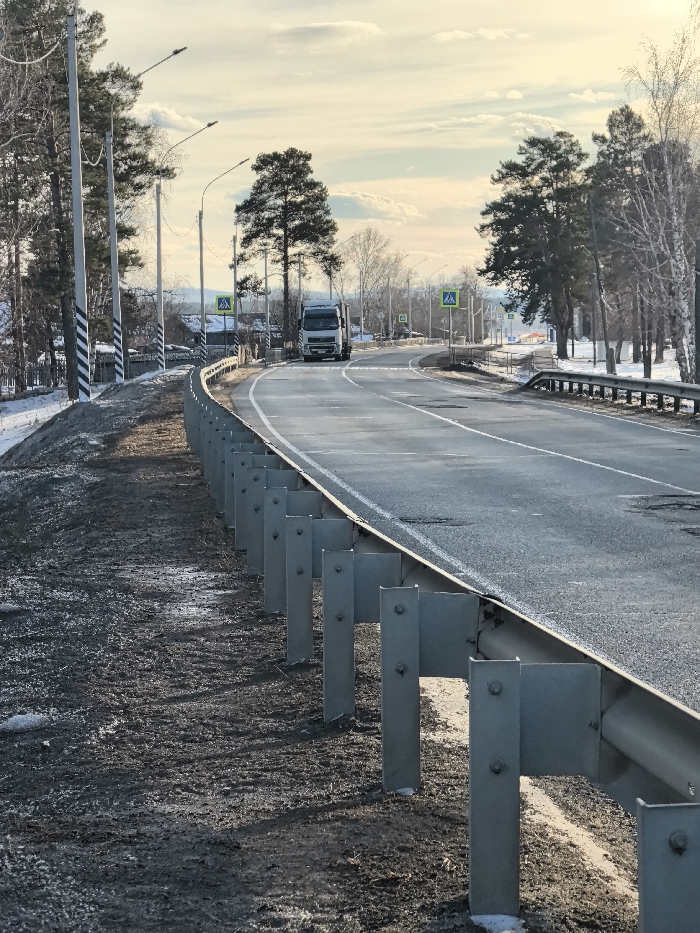The image displays a two-lane asphalt road curving gently to the right in Аблатуйский Бор, Russia, under an overcast sky. A galvanized steel guardrail lines the left side of the road, separating it from an unpaved shoulder of dark soil, mud, and melting snow patches. Several utility poles with streetlights are positioned along the left shoulder, some featuring black and white striped bases. In the distance, a white semi-truck without a visible trailer is observed traveling away from the viewer on the right lane of the curved road. Multiple yellow triangular pedestrian crossing signs are visible along both sides of the road in the background. The environment is characterized by a mix of evergreen and bare deciduous trees lining both sides of the road, with small structures visible amongst the trees on the left. The asphalt pavement shows signs of wear, including cracks and patches. No people are visible.
