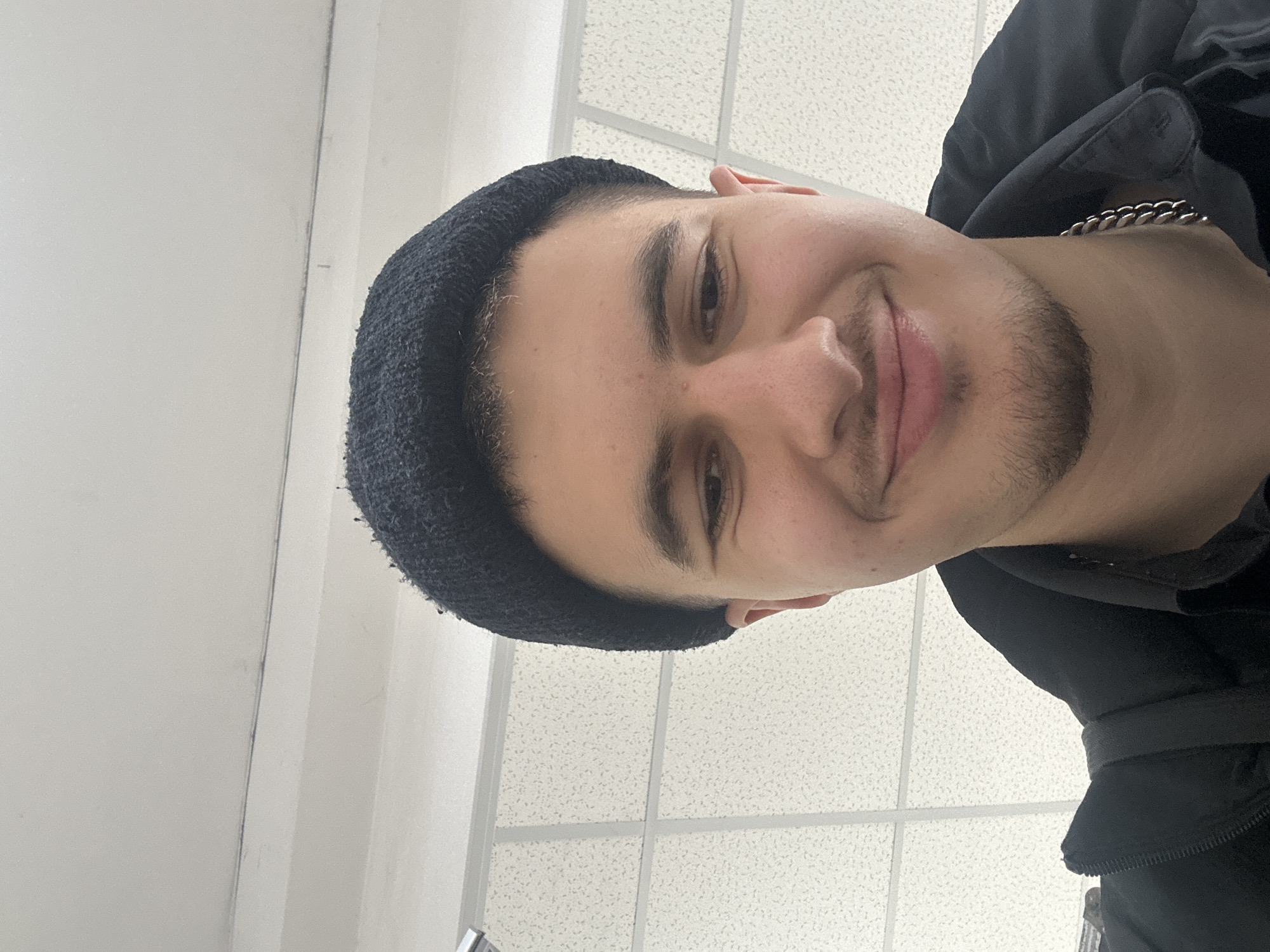 A selfie of a young man wearing a black beanie, black jacket, and a silver chain necklace. The background shows a white tiled ceiling and a white wall. Location cannot be determined from the image.