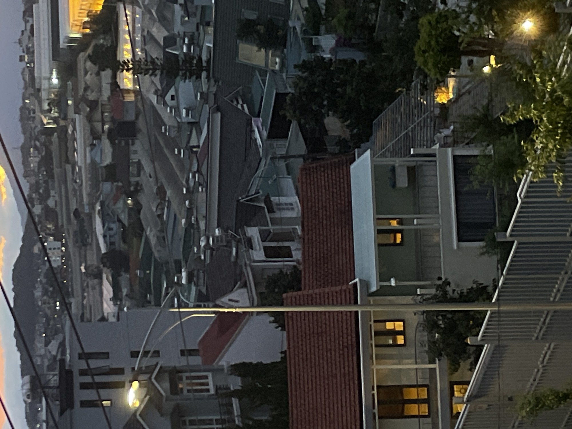 The image depicts a twilight view of a hillside city, likely Lâm Viên - Đà Lạt, Vietnam, characterized by a dense collection of houses and buildings with varying roof designs, predominantly red and dark grey tiles. The architecture suggests a mix of residential and commercial structures, some appearing to be modern with balconies and large windows, while others have more traditional pitched roofs. Numerous greenhouses or similar agricultural structures are visible across the landscape, indicating a possible agricultural hub. The lighting in the scene is a mix of natural twilight and artificial illumination emanating from windows, streetlights, and possibly illuminated buildings in the distance, creating a warm glow against the darkening sky. Several trees and dense foliage are interspersed among the buildings, adding greenery to the urban environment. A metallic fence runs along the foreground, with some vegetation growing near it. The overall impression is of a sprawling, settled urban area on a mountainous terrain, with signs of ongoing activity as evening approaches.